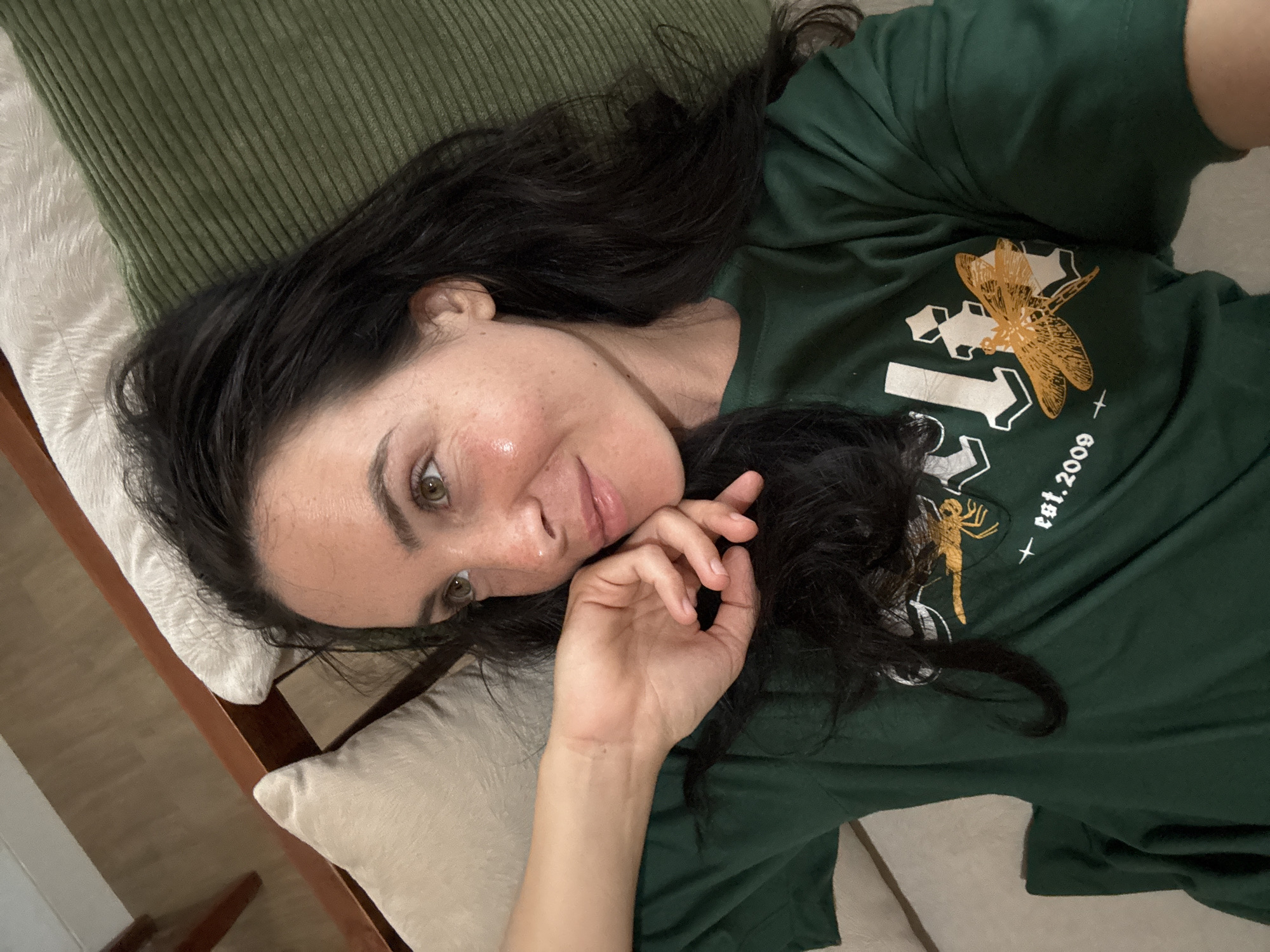 A woman is lying on a couch with a green pillow behind her head. She is wearing a dark green t-shirt with a graphic design that includes a dragonfly and the text "est. 2009". Her left hand is resting on her cheek, and she is looking to her right. The background consists of a wooden floor and part of a wooden chair.