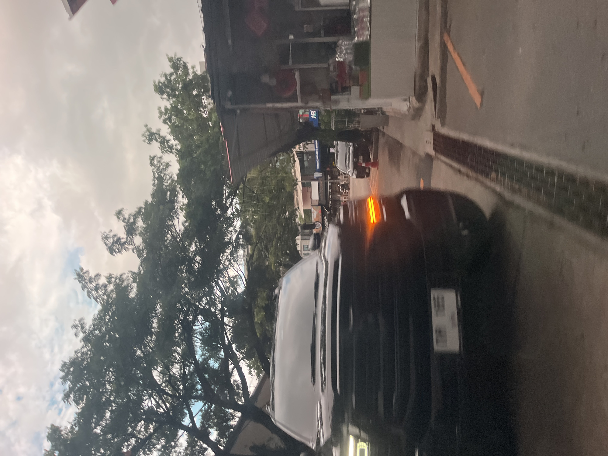 The image shows a dark-colored SUV parked on a street in Choeng Thale, Thailand. The vehicle is positioned on the right side of the frame, with its front facing towards the viewer. The SUV has its headlights on, emitting a bright white light. The license plate is partially visible and appears to have Thai characters and numbers.
In the background, there are several buildings with awnings, suggesting a commercial or market area. Trees with lush green foliage line the street, and the sky is overcast with clouds. A drainage grate is visible on the road in front of the SUV. To the right of the SUV, a small building with a covered area displays shelves with various items, possibly for sale. The overall scene suggests a typical street view in a bustling area of Choeng Thale.