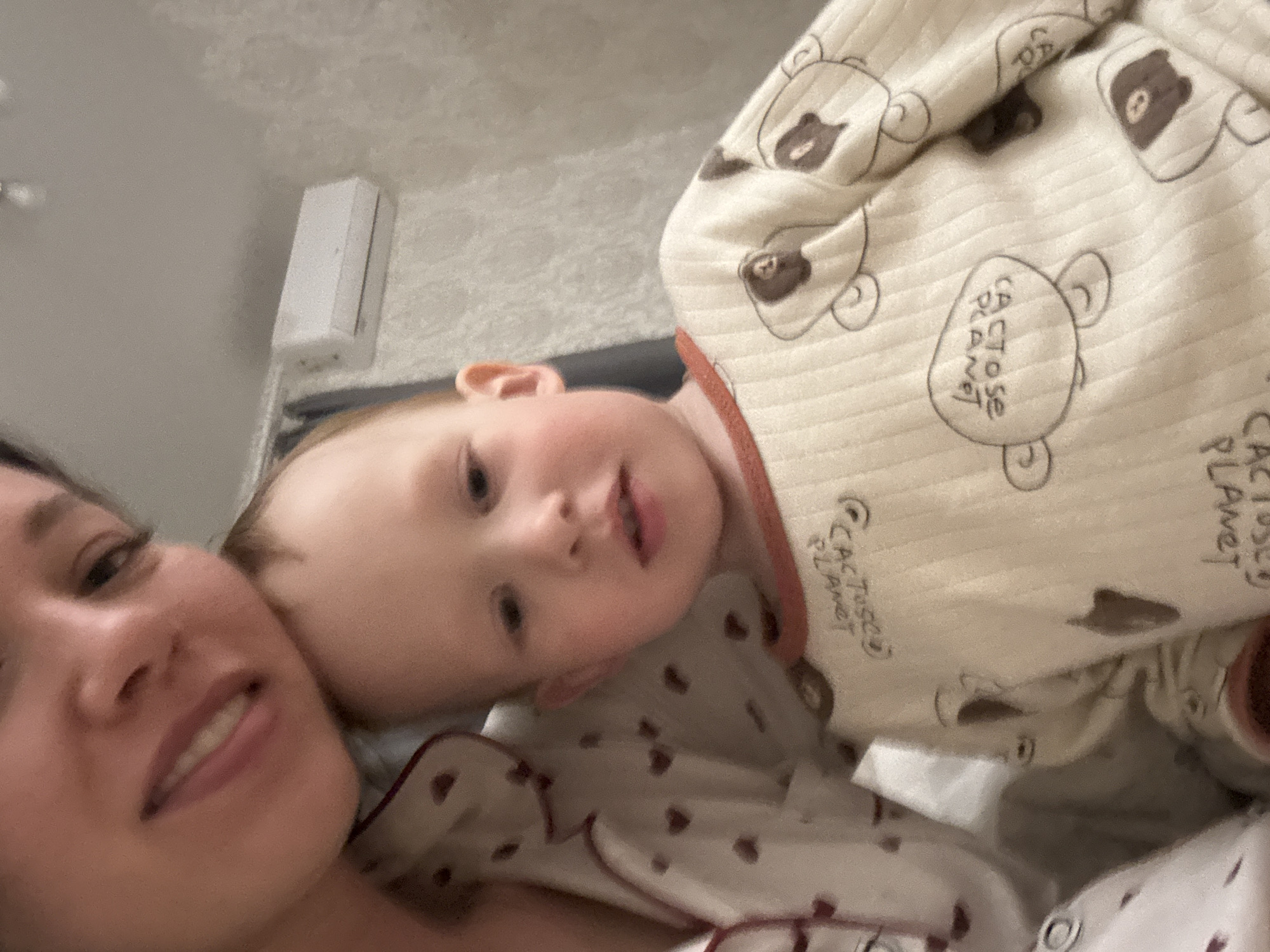 A close-up shot depicts a woman and a baby. The woman's face is in the lower left corner, showing part of her forehead, eyes, nose, mouth, and chin. The baby is lying near her, with their face turned towards the camera. The baby is wearing pajamas with a "Cactus Planet" pattern. A white air conditioner is visible in the upper left background, mounted on the wall. The location is Ферма, Russia.