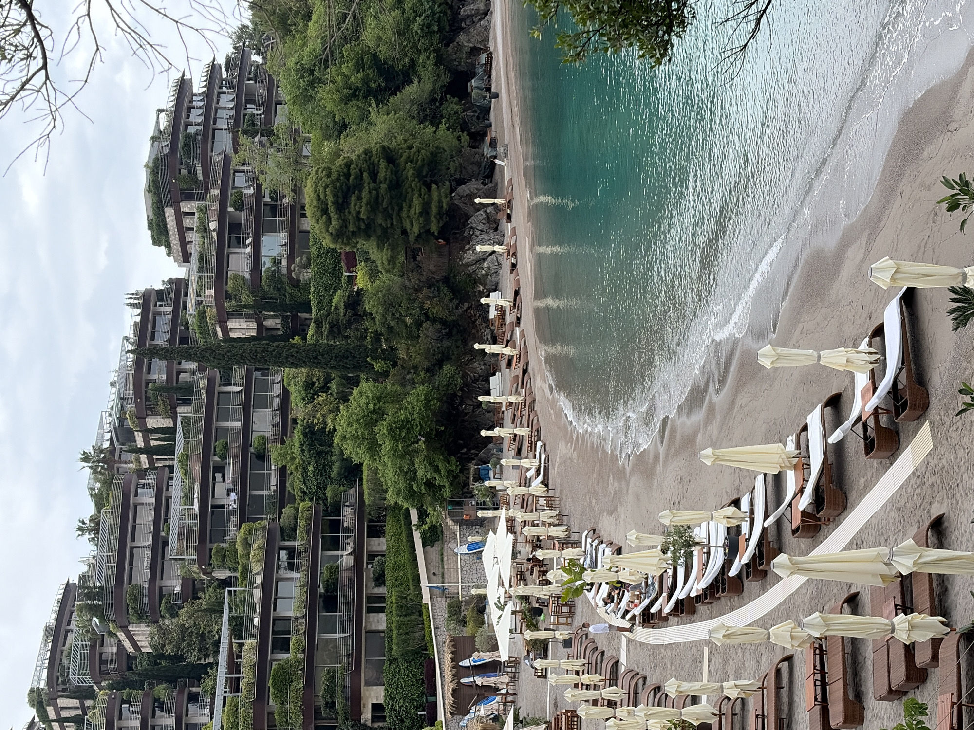 The image depicts a view of a beach in Budva, Montenegro. The beach has rows of sun loungers with white umbrellas, arranged in an orderly fashion along the sand. Turquoise sea waves are visible along the shoreline. To the left, a modern apartment building is visible. The building is covered in greenery. The sky is overcast, suggesting a cloudy day.
