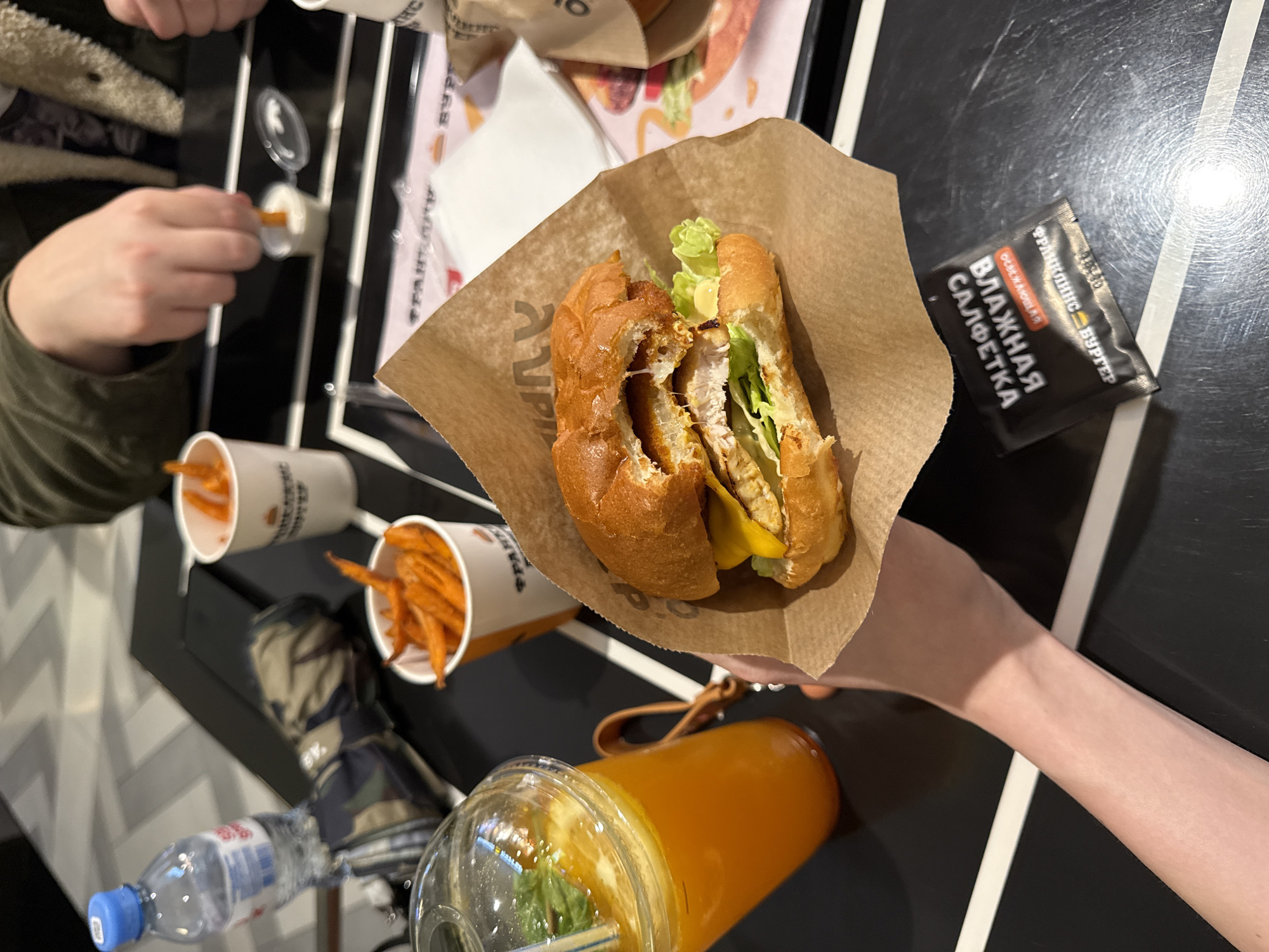 A close-up, top-down view shows a person holding a partially eaten burger in a brown paper wrapper. The burger has a golden-brown bun, melted cheese, a crispy chicken patty, and lettuce. To the left, a clear plastic cup contains an orange beverage with mint and a straw. In the background, two white paper cups are filled with french fries, and one person's hands are visible, with one hand dipping a fry into a small container of sauce. A bottle of water with a blue cap is also present. The setting appears to be a casual dining establishment, likely a fast-food restaurant, with a black table surface marked with white lines. The restaurant's logo, featuring Cyrillic script, is visible on the burger wrapper and the french fry cups, and based on context from similar images, this establishment is likely "Burger Heroes" located in Khimki, Russia. The overall scene depicts a meal being consumed.