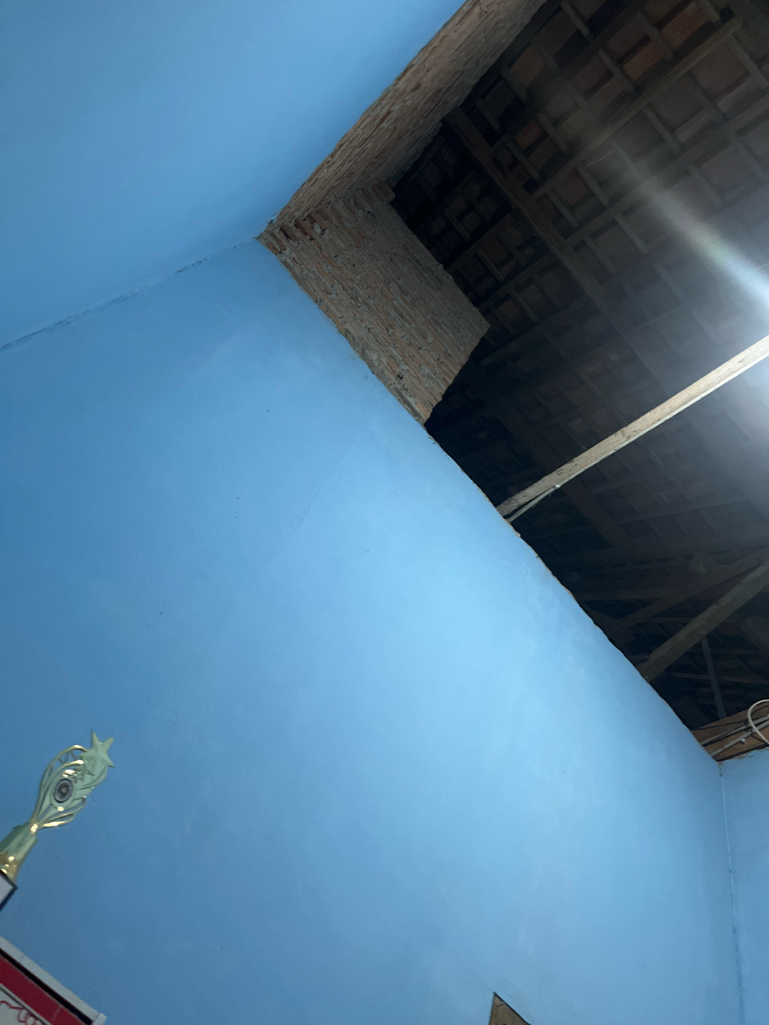 This is an interior shot of a room, likely a residential space, in Kebumen, Indonesia. The walls are painted a light blue. In the foreground, on the left, a golden trophy with a star on top is visible. The ceiling is unfinished, revealing exposed wooden beams and terracotta tiles, suggesting ongoing construction or renovation. A bright light source, possibly a lamp or sunlight, is illuminating the scene from the upper right, creating a lens flare effect. The perspective is from a low angle, looking upwards towards the corner where the walls meet the ceiling.