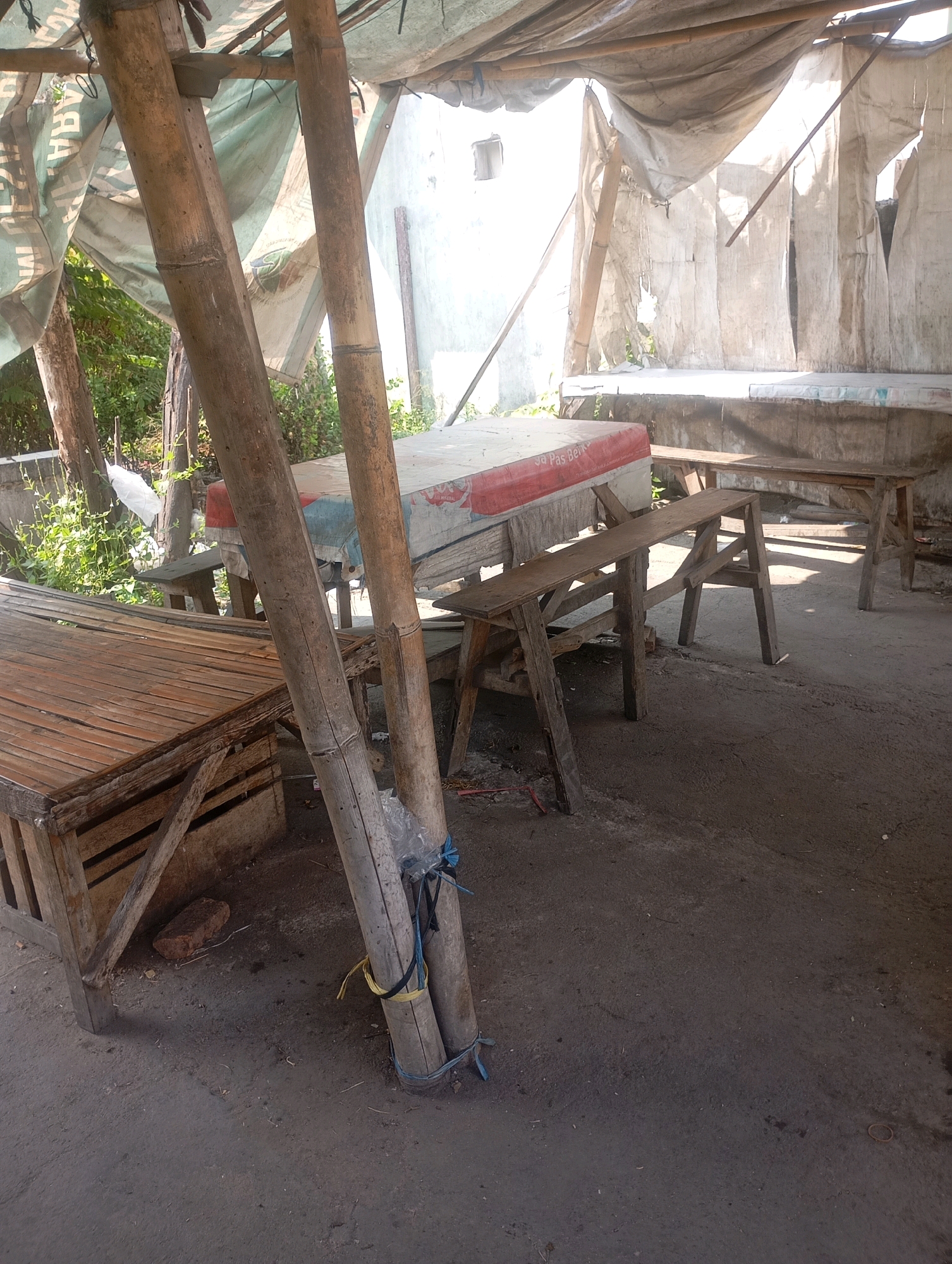 In Babelan, Indonesia, an outdoor seating area is depicted with simple, rustic furniture. The space is partially covered by a makeshift roof constructed from canvas or tarp supported by bamboo poles. Wooden tables and benches, some made from bamboo, are arranged on a dirt or concrete ground. One table has a mattress on it, suggesting it might be used for lounging or perhaps even sleeping. The environment appears to be outdoors with some vegetation visible in the background. The overall scene conveys a sense of a casual, unpretentious gathering spot.