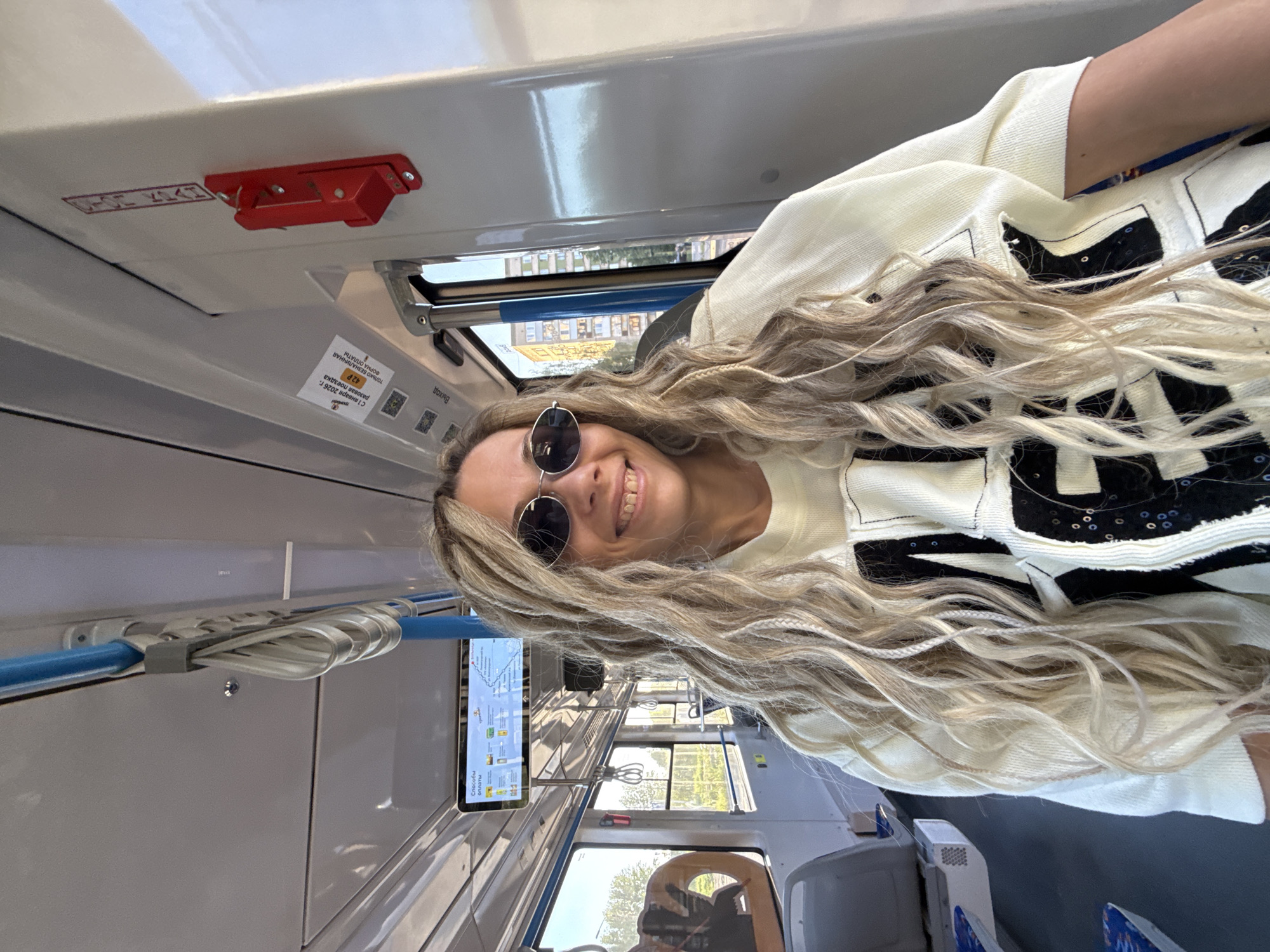 A female with long, blonde, wavy hair, including some braided sections, is seated inside a public transport vehicle, smiling directly at the camera. She wears round dark sunglasses and a light-colored, long-sleeved top with large black irregular patches, some adorned with small sequins. The vehicle's interior features white and grey panels, a blue overhead grab bar with white hanging straps, and large windows. Through the windows, an urban landscape with trees and multi-story buildings, including one with blue and white sections, is visible under daylight. Overhead, a digital display shows "Способы оплаты" (Methods of payment) and what appears to be a route map. Below the display, a sticker indicates "С 1 января 2023 г. разовая поездка 42 Р." (From January 1, 2023, a single trip is 42 R.) and "только безналичная форма оплаты" (cashless payment only). A red emergency lever mechanism is mounted on the wall to the right. A sign displaying "Выход" (Exit) is also visible. The scene takes place within a public transport vehicle in the city of Rusanovo, Russia.