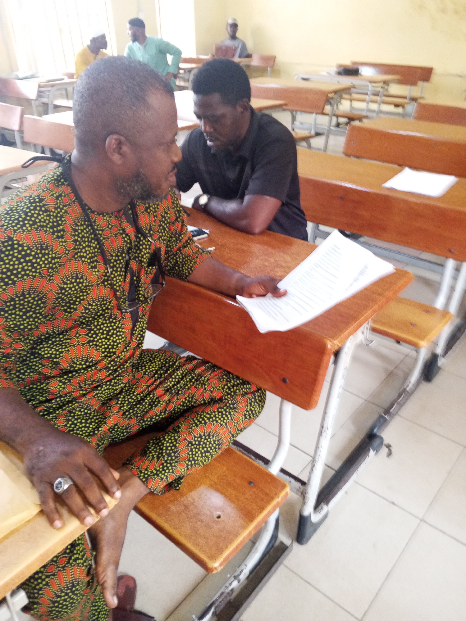 The image depicts an indoor scene, likely a classroom or lecture hall, in Lokoja, Nigeria. In the foreground, a man wearing a vibrantly patterned dashiki with green, yellow, and red motifs sits at a wooden desk. He has a beard and is wearing a ring on his left hand. He is holding a document with text on it. To his right, another man in a black t-shirt is seated, looking down. The desks are made of wood and metal, typical of educational institutions. In the background, other individuals are visible, some standing and others seated at desks, suggesting a group gathering. The room has light-colored walls and tiled flooring. The overall atmosphere appears to be one of an educational or a formal meeting setting.