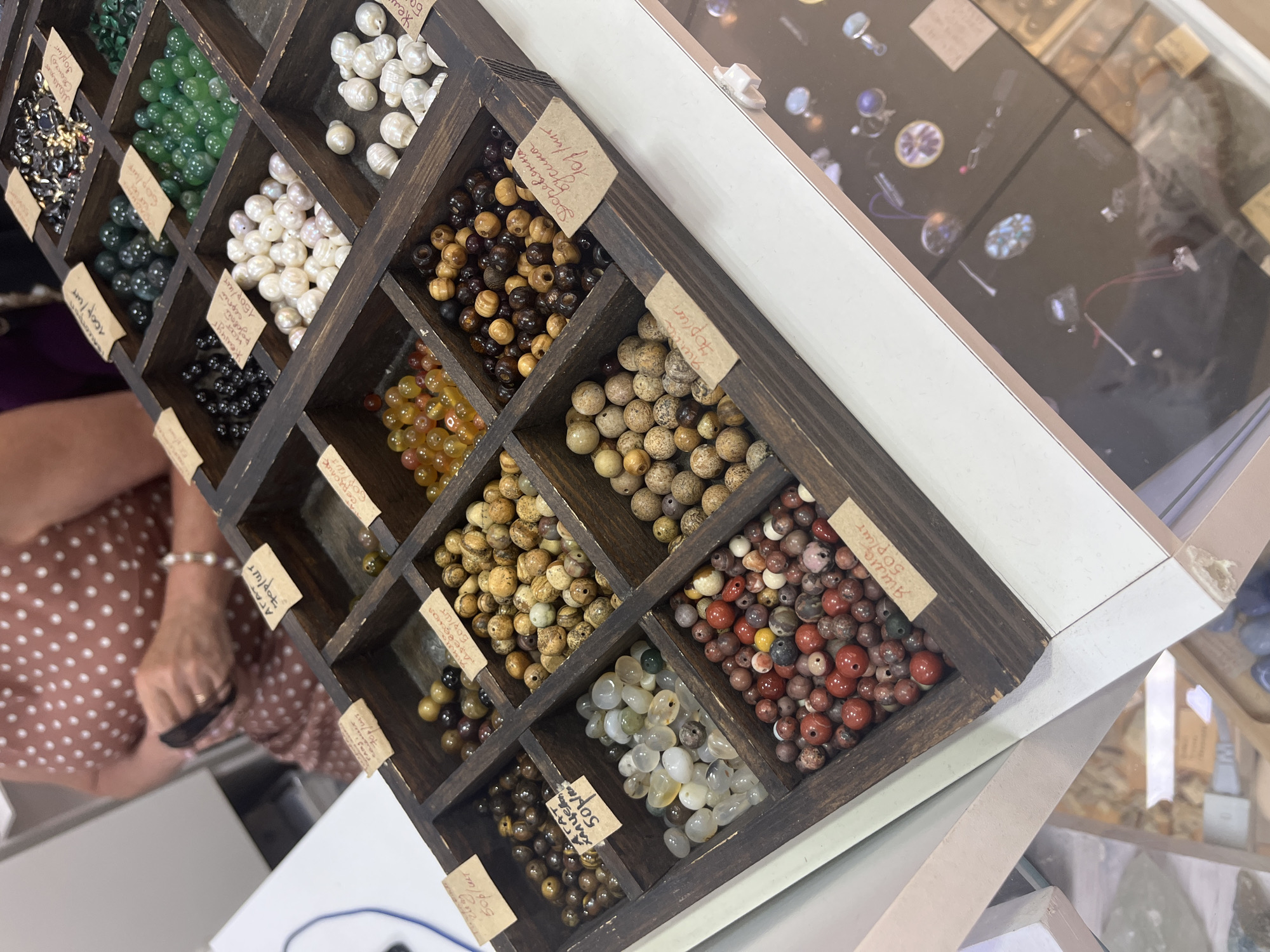 This is a close-up, eye-level shot of a wooden display box filled with various types of beads, likely at a market or craft fair. The box is divided into multiple compartments, each containing a different type of bead. Some compartments contain small, dark, possibly obsidian or jet beads, while others hold larger, milky white pearls. There are also compartments with green, jade-like beads, and others with amber-colored beads. Several compartments are filled with natural-toned, spotted, and banded beads, appearing to be agate or jasper. One compartment contains small, reddish-brown and grey beads. Small, handwritten paper tags with prices in rubles are tucked into some of the compartments. In the background, a person wearing a brown polka-dot dress is partially visible, their hands occupied with something unseen. The overall scene suggests a retail environment specializing in jewelry-making supplies. The image does not contain any specific visual cues to pinpoint the location to Sochi, Russia.