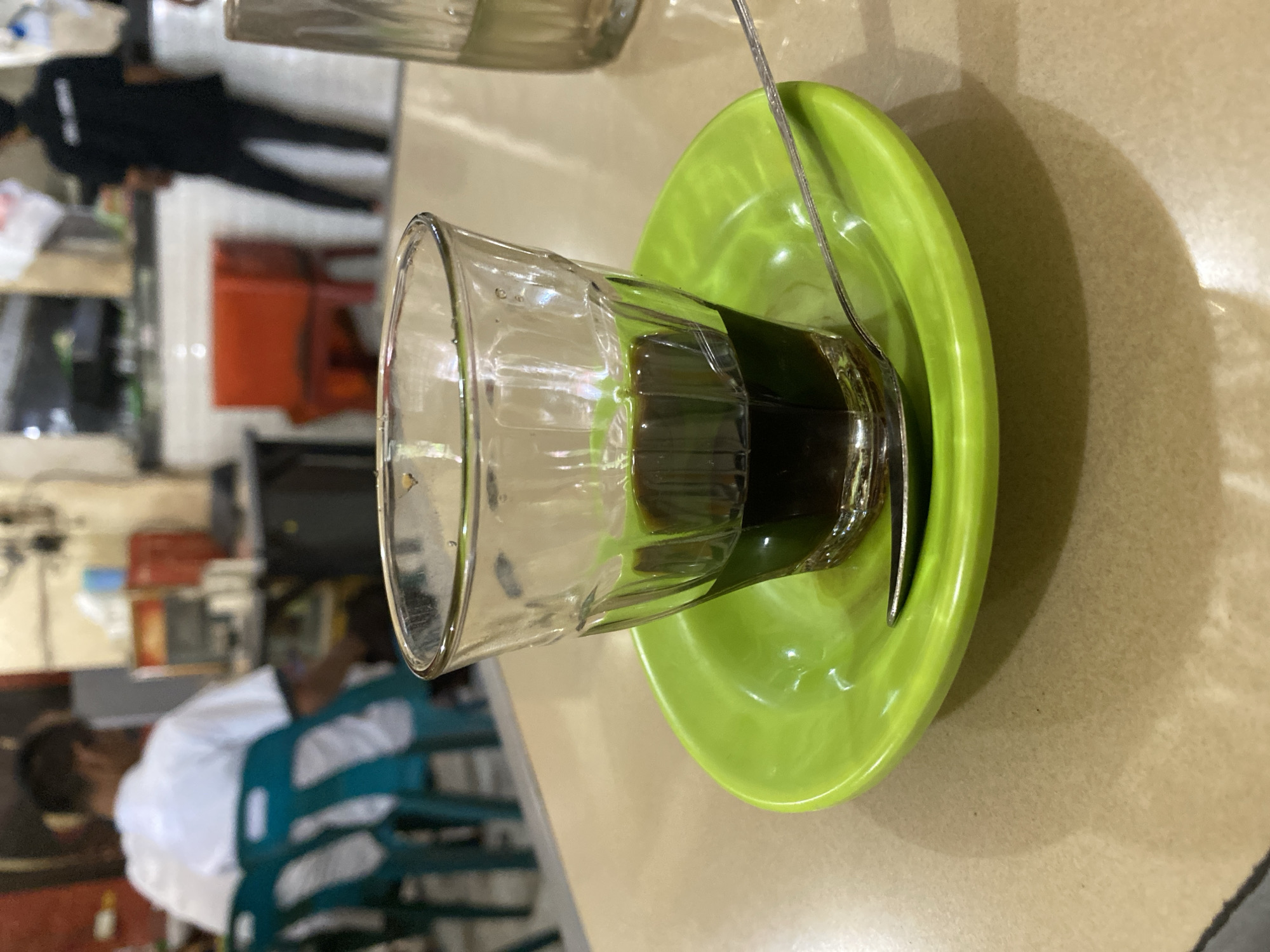The image depicts a close-up of a small glass of dark liquid, likely coffee, sitting on a bright green saucer. A silver spoon rests on the saucer, partially submerged in the liquid. The setting appears to be an indoor establishment, possibly a cafe or restaurant, given the presence of tables, chairs, and people in the background. In the blurred background, a person wearing a white shirt and another in a black shirt are visible. The environment suggests a casual, everyday setting, common in urban areas like Lhokseumawe, Indonesia. The lighting suggests it is daytime, possibly indoors with artificial lighting contributing to the overall ambiance.
