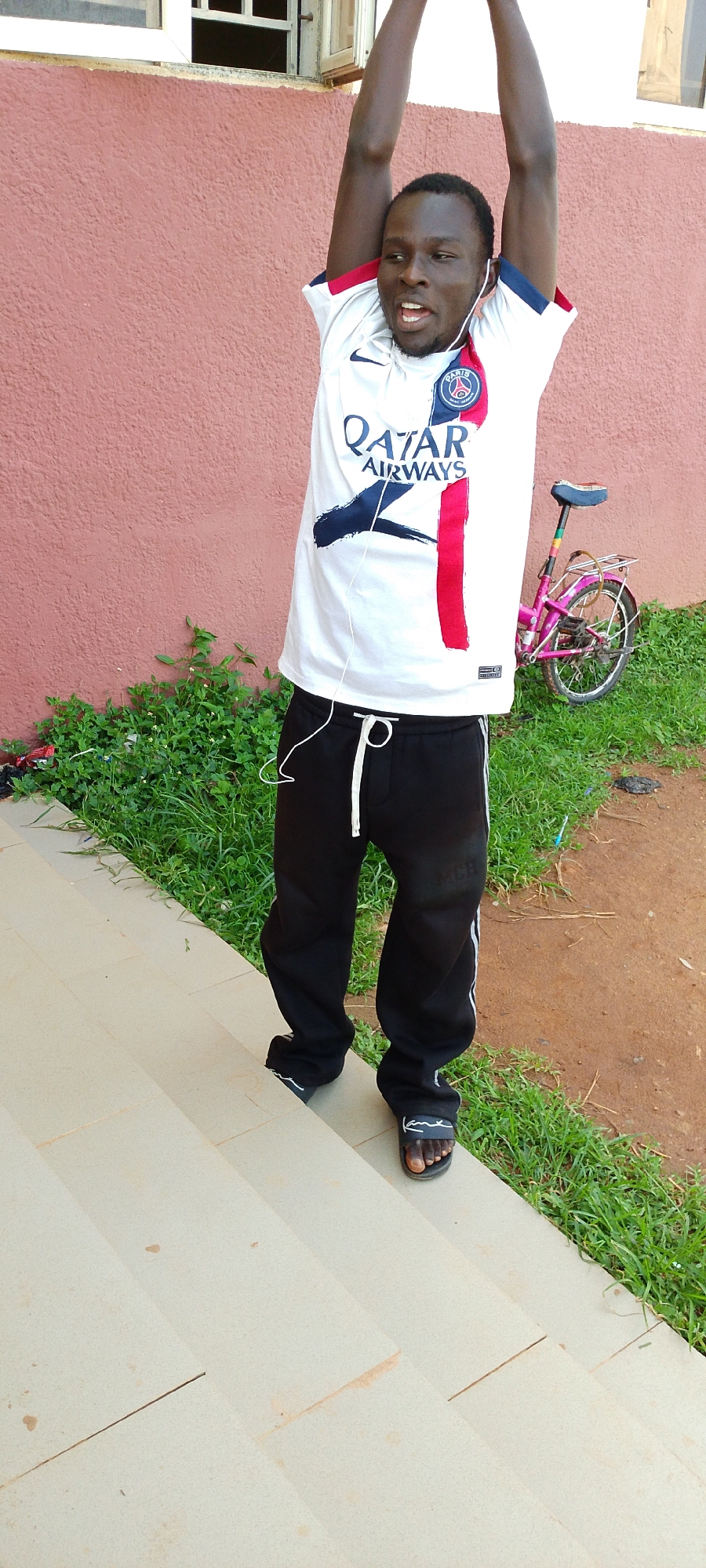 A young Black male stands outdoors on a concrete stairway, facing forward with his arms raised overhead. He is wearing a white jersey with "Qatar Airways" emblazoned across the chest, black sweatpants with white stripes down the sides, and black slide sandals. White earbuds are visible in his ears, with the cord hanging down the front of his jersey. The man is looking slightly to his left with his mouth open as if speaking or yawning. Behind him, a pink bicycle with a blue seat and a rear rack is leaning against a textured, pink wall. Lush green grass and a dirt patch are visible to the right of the stairway and behind the bicycle. The scene appears to be in a residential area in Gumel, Nigeria, likely in a courtyard or near the entrance of a building. The overall lighting suggests it is daytime, possibly late morning or early afternoon, with diffused sunlight.
