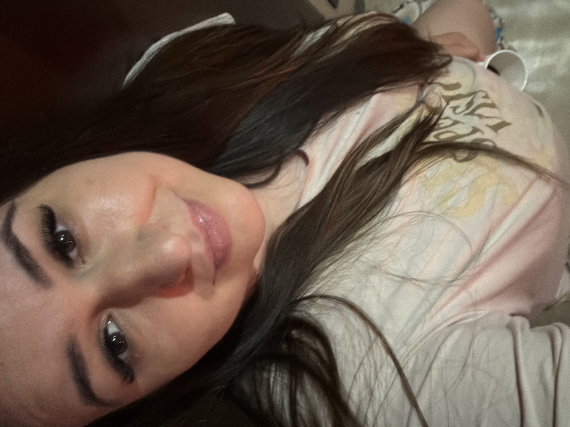 A selfie of a woman with long dark hair is shown. She is wearing a light-colored t-shirt with a graphic print. Her eyes are dark and she has long eyelashes. She is smiling and her lips are glossy. The background is out of focus and appears to be an indoor setting.