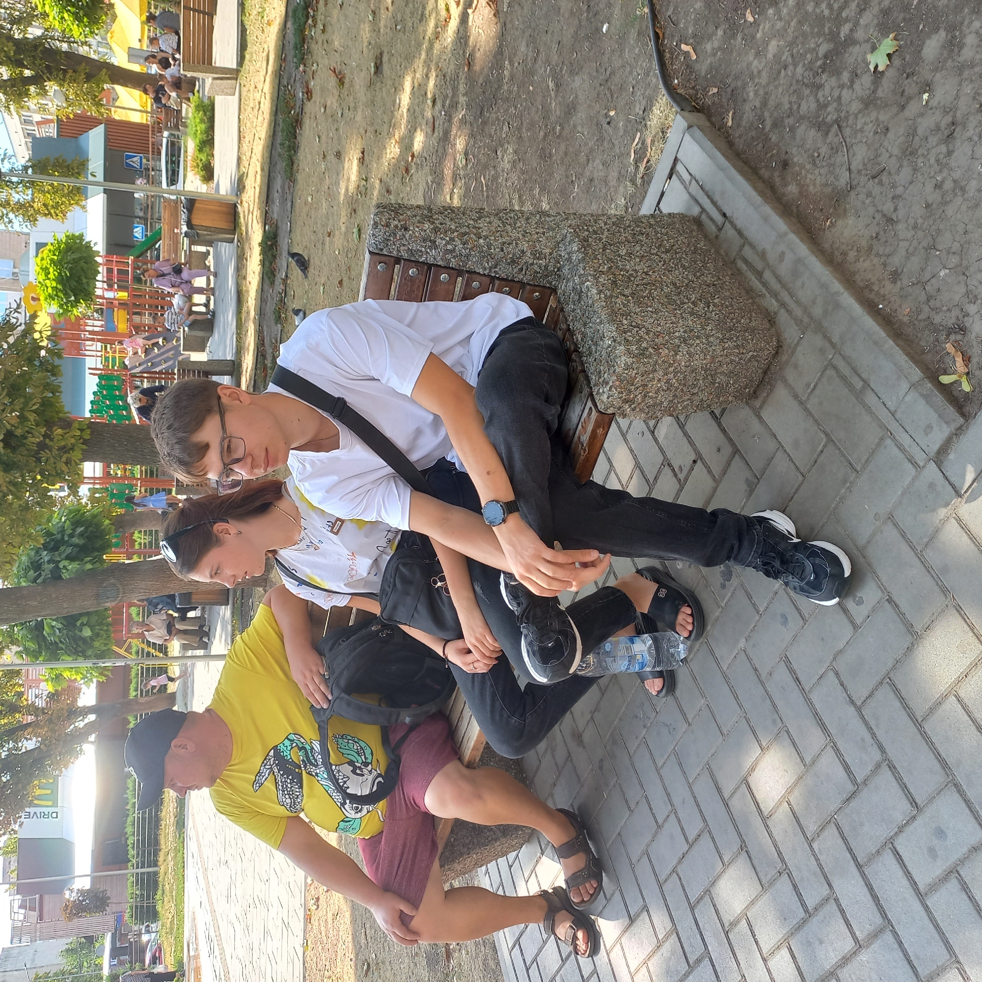The image shows a street scene in Kropyvnytskyi, Ukraine, on a sunny day. Three people are sitting on a bench in a park-like setting. The person on the left, a man, wears a yellow t-shirt with a graphic print and dark shorts. Next to him sits a young woman wearing a white t-shirt and black jeans, with sunglasses on her head. To her right is a young man in a white t-shirt and black jeans, wearing glasses and a watch. A black backpack is placed on the bench behind the woman and young man. In the foreground, a plastic bottle of water is on the ground. The background shows trees, a playground with colorful equipment, and some buildings, one with a "DRIVE" sign. The ground is paved with interlocking bricks, and dirt with scattered leaves is visible beyond the paved area.