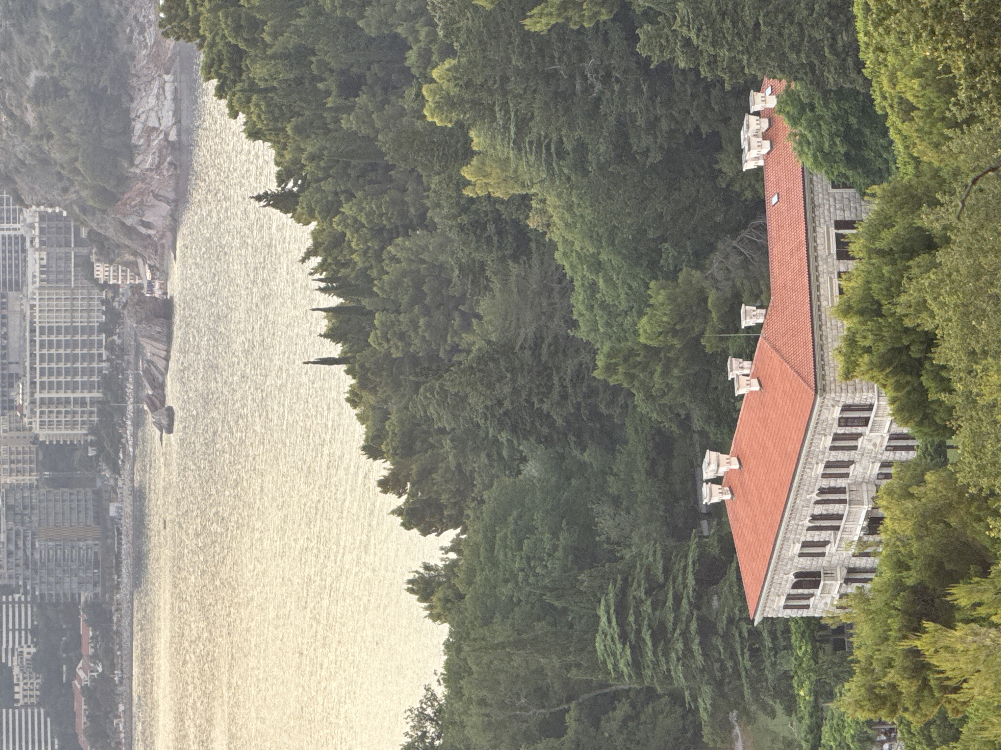 The image depicts a serene coastal scene with a notable building and lush greenery. The primary subject is a long, multi-story building with a red tiled roof and stone facade, featuring numerous windows and decorative architectural elements. This structure is nestled within a dense forest of tall, green trees that extend across the frame.
To the left of the building and forest, a body of water, likely the Adriatic Sea, stretches out. The water is calm and reflects the soft light, suggesting it is either early morning or late afternoon. Along the coastline, a town or city with many buildings is visible, characterized by a grid-like pattern of structures. The buildings are clustered closely together, forming a dense urban landscape that contrasts with the natural beauty of the surrounding trees and the sea.
In the distance, a rocky cliff face is visible, adding to the dramatic topography of the location. The overall impression is one of a picturesque setting where human habitation coexists with a vibrant natural environment. The lighting and the gentle shimmer on the water suggest a peaceful atmosphere. No specific activity or event is clearly depicted, and no text is visible in the image. The scene appears to be a landscape view, possibly taken from an elevated vantage point.