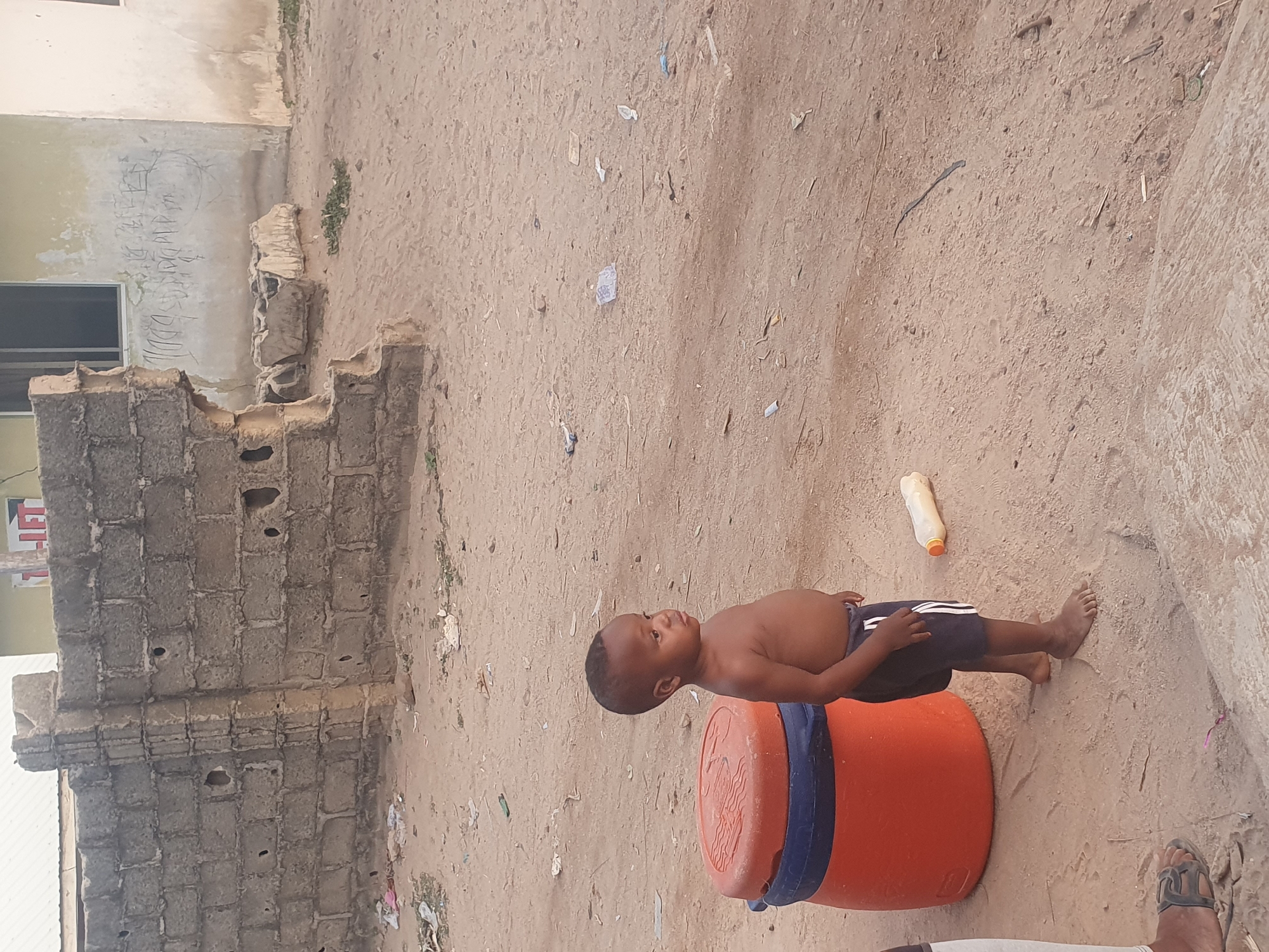 The scene depicts a young, shirtless child standing beside an orange barrel with a blue stripe, in an outdoor setting in Lokoja, Nigeria. The child is dark-skinned and has short hair. He is wearing dark-colored shorts. His bare feet are on the sandy ground. Behind the child, a plastic bottle lies horizontally. The background is sandy and littered with debris. To the left, there is a concrete block structure, and a building with visible text on the wall. A foot wearing a sandal is partially visible in the lower part of the image.