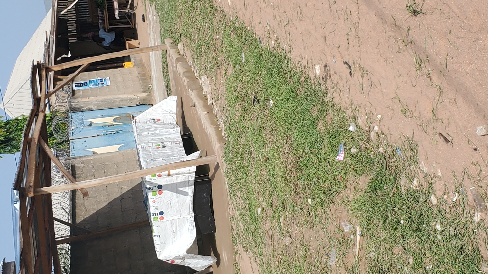 The image depicts an outdoor scene in Jalingo, Nigeria, focusing on a covered structure with a white tarp draped over a table-like surface. The structure has a corrugated metal roof supported by wooden posts. Behind the structure, a brick wall with barbed wire is visible, along with a blue metal gate. To the right of the gate, a small building with a white roof is present, and a person is seated inside on a bench. The foreground of the image shows a dirt and grass area littered with debris, including plastic bottles and wrappers. The lighting suggests it is daytime, with shadows cast by the covered structure.