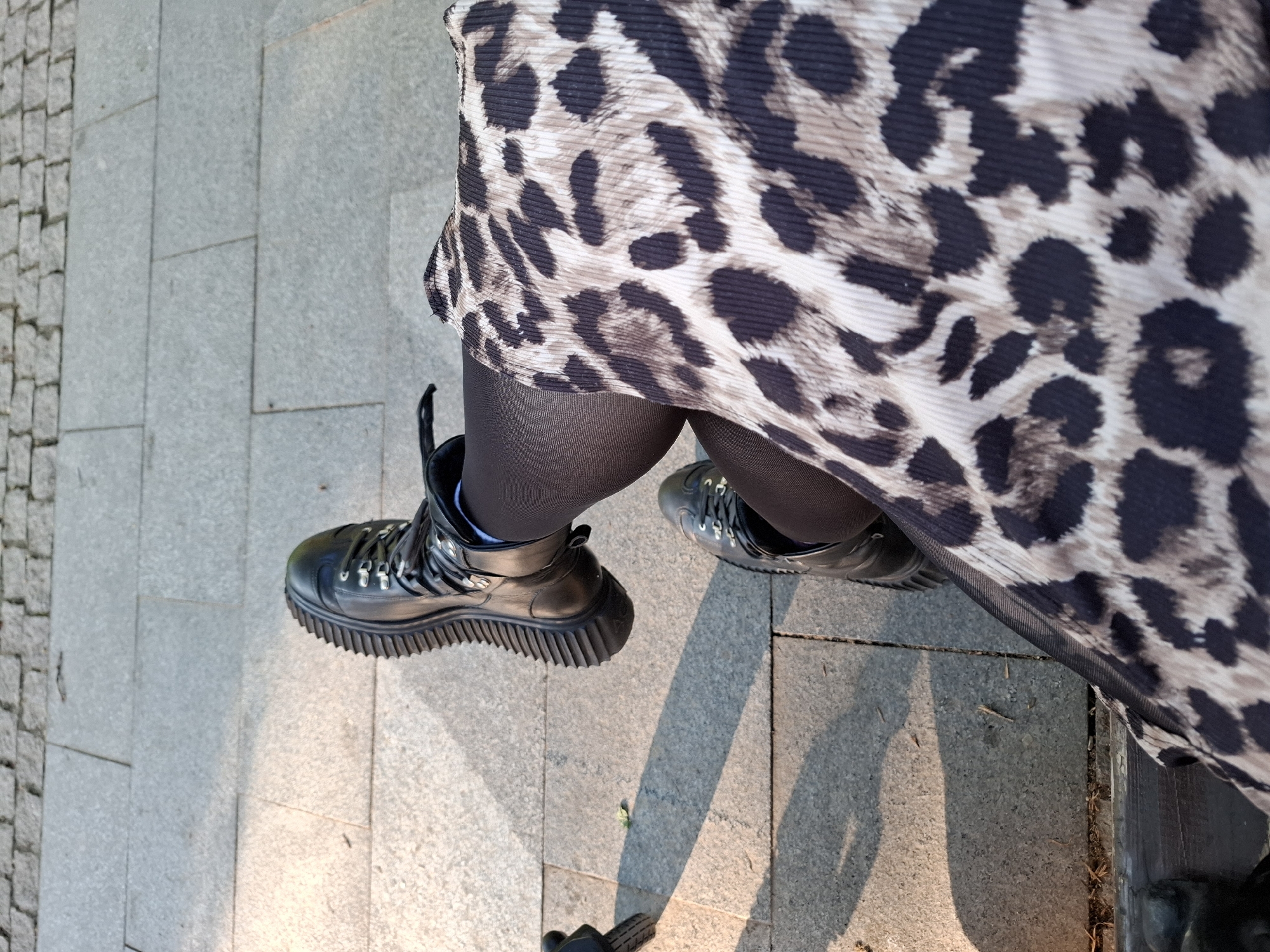 A high-angle shot shows a person standing on a paved surface, possibly a sidewalk or public space in Novi Sad, Serbia, based on the style of the paving stones. The person is wearing black tights, black boots with a thick, ridged sole, and a garment with a leopard-print pattern. The lower part of the person's body and feet are primarily visible, with the leopard-print garment obscuring the rest of the body. The pavement consists of large, gray square tiles, alongside a section paved with smaller, rectangular stones. Sunlight creates shadows on the pavement.