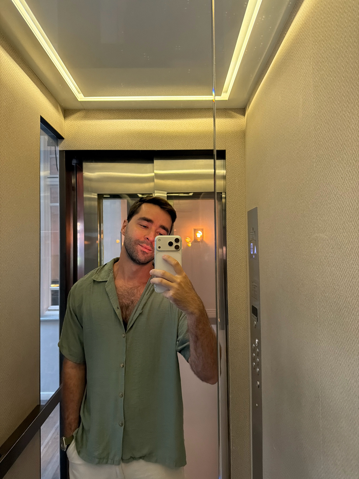 A man is taking a selfie in an elevator in Tel Aviv, Israel. He is wearing a green short-sleeved shirt and white pants, with his left hand in his pocket. The elevator walls are textured beige wallpaper, and the ceiling has recessed LED lighting. The elevator doors are stainless steel, and a control panel with illuminated buttons is visible on the right wall. The man is looking at his phone, which is held in his right hand. The reflection in the elevator doors shows a window with buildings outside, suggesting an urban environment.