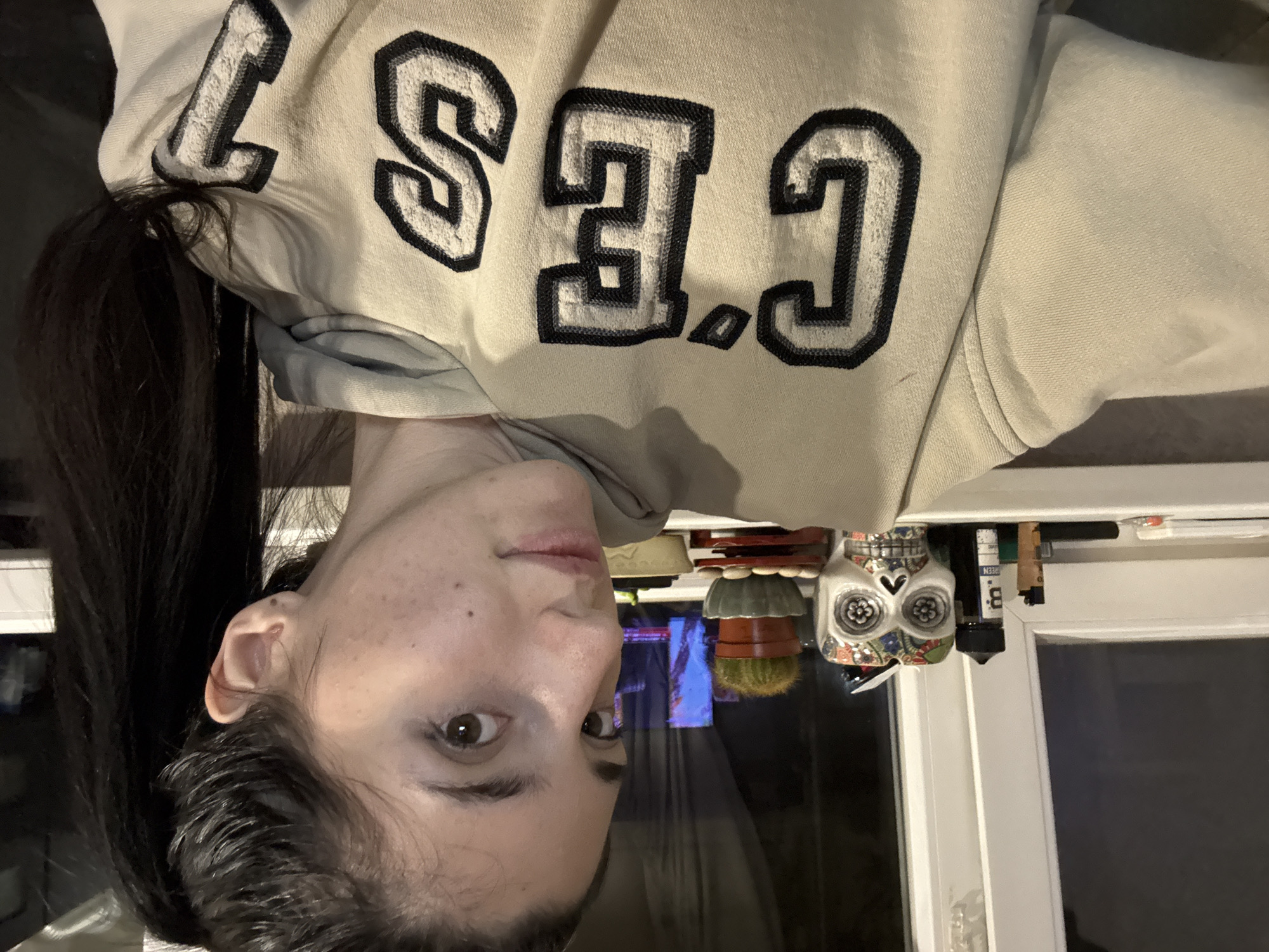 A woman is wearing a "CES" lettered sweater. She is indoors, with items, including a decorative skull and stacked pots, visible in the background. The location is indeterminable without further information.
