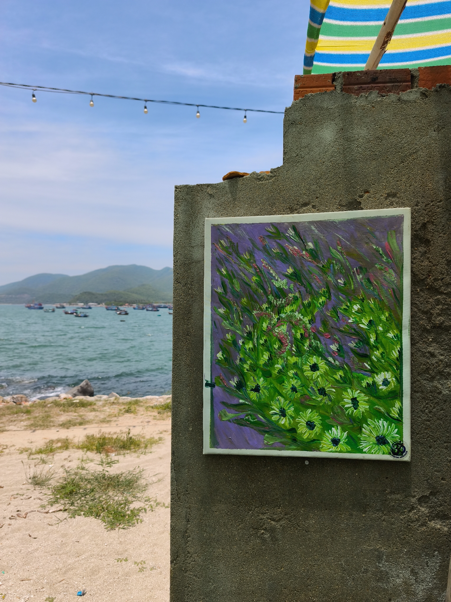 The image depicts an outdoor scene in Vĩnh Lương, Vietnam, on a bright, clear day. A grey concrete pillar occupies the right side of the frame, mounted with a square painting of abstract green and purple flowers. The painting is bordered by a white frame. To the left of the pillar, a sandy beach slopes down to the sea. The water is a vibrant turquoise, with several small boats anchored offshore and a hazy mountain range in the background. Above the beach, a strand of white string lights is suspended against a pale blue sky with thin, wispy clouds. A colorful striped awning, made of yellow, green, and blue fabric, is visible at the top right of the image, attached to a brick structure. The overall impression is one of a casual, seaside setting, possibly a cafe or an artist's workspace, showcasing local art against a natural backdrop.