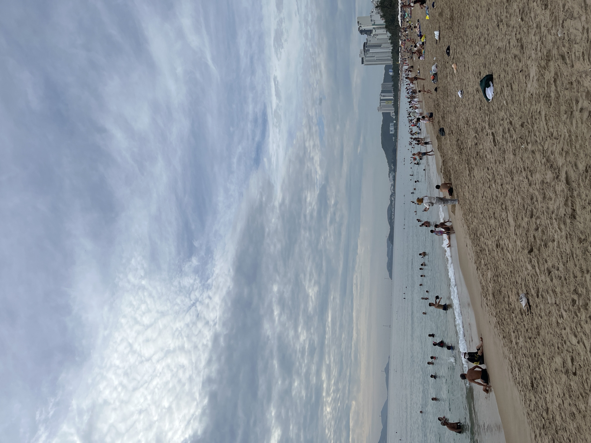 The image captures a cloudy day at a crowded beach in Nha Trang, Vietnam, likely a section of the main city beach. The sand in the foreground is light brown and textured, with several scattered items like a pair of white sneakers, a dark green towel, and other unidentifiable objects. A few people are sitting or lying on the sand closer to the water's edge. The waterline is marked by small waves breaking on the shore.
The ocean is populated with numerous people, some wading, others swimming, and a few further out in the water. The water is a muted gray-blue, reflecting the overcast sky. In the distance, the shoreline is lined with buildings, including several tall, modern high-rise hotels and apartments, characteristic of Nha Trang's urban coastal development. Hills can be seen further inland, and the outline of islands or peninsulas is visible on the horizon. The sky is filled with a complex pattern of gray and white clouds, creating a dramatic, diffused light.