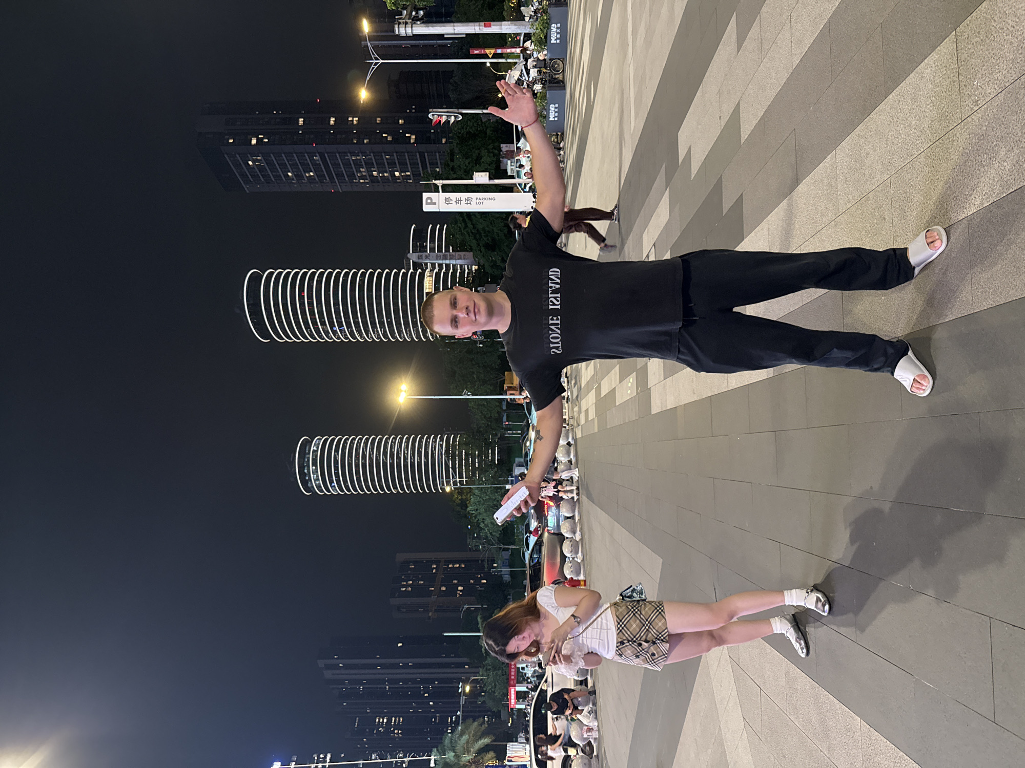 In Tianhe District, China, at night, a Caucasian man in a black t-shirt with "STONE ISLAND" printed on it and black sweatpants stands with his arms outstretched. Beside him, a young East Asian woman in a white top and plaid skirt looks at her phone. The background shows modern skyscrapers, including two distinctive cylindrical buildings with white ring lighting, and a street with cars and pedestrians. The ground is paved with large rectangular tiles.