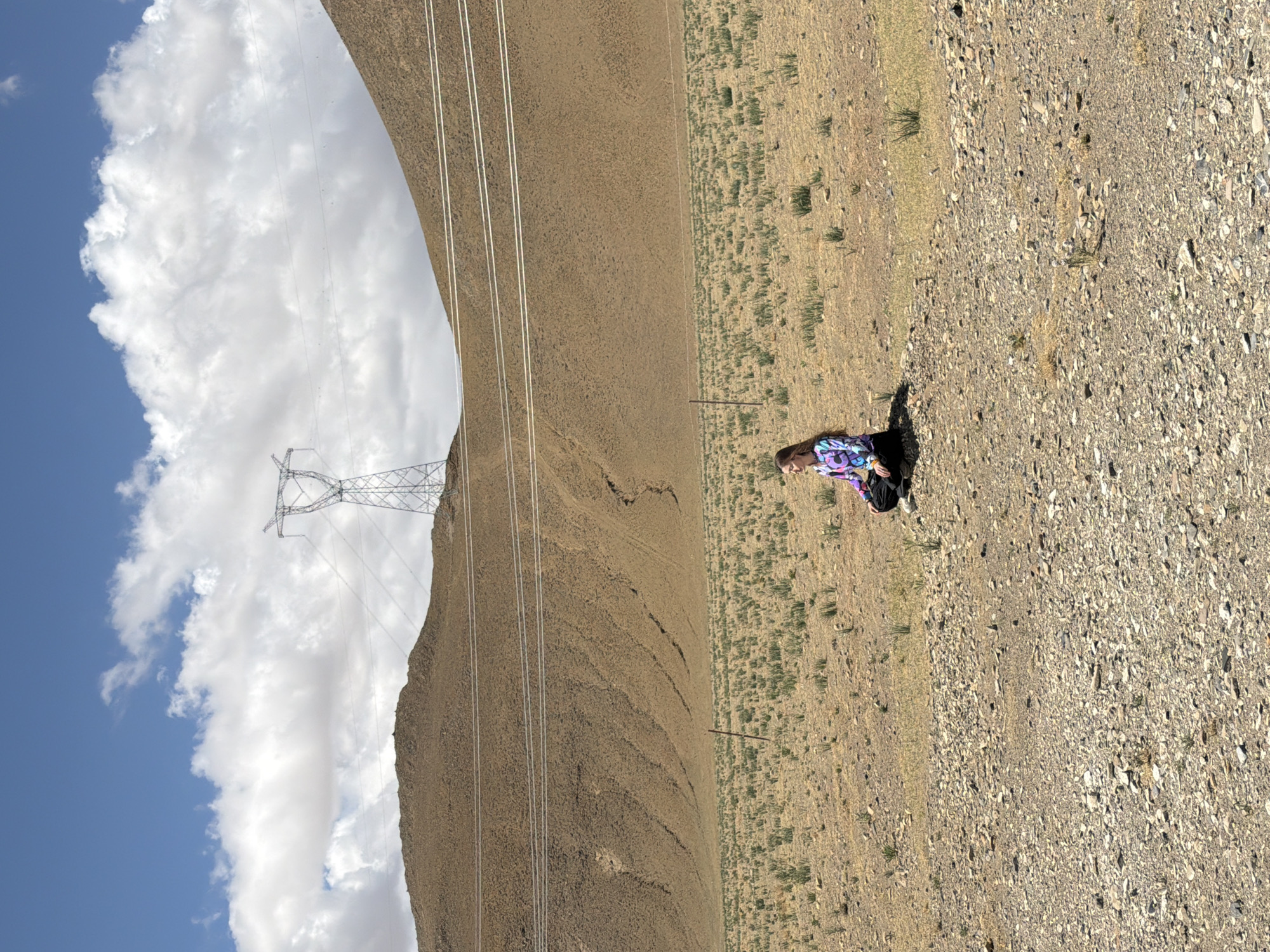 The image, originally oriented sideways, displays a vast, arid outdoor landscape under a bright, partly cloudy sky. If rotated to its correct horizontal alignment, the scene would show a wide expanse of dry, rocky ground in the foreground and middle ground, with sparse vegetation and small clumps of dry grass.
On the right side of the frame, a person with blonde or light brown hair is seated or kneeling on the ground, wearing a colorful, long-sleeved shirt and dark pants, with a black backpack resting beside them. Their gaze is directed downwards.
To the left, a large metal electricity transmission tower stands, with multiple power lines extending horizontally across the mid-ground. Behind the pylon and power lines, a prominent, barren mountain range stretches across the left half of the image, its slopes showing visible texture and lines. The sky above is a clear blue, adorned with large, fluffy white cumulus clouds. The bright sunlight indicates it is daytime, casting clear shadows.
No text is visible in the image. The scene evokes a sense of vastness and solitude in a remote, possibly high-altitude environment, consistent with the provided location context of Mainpu township, China.