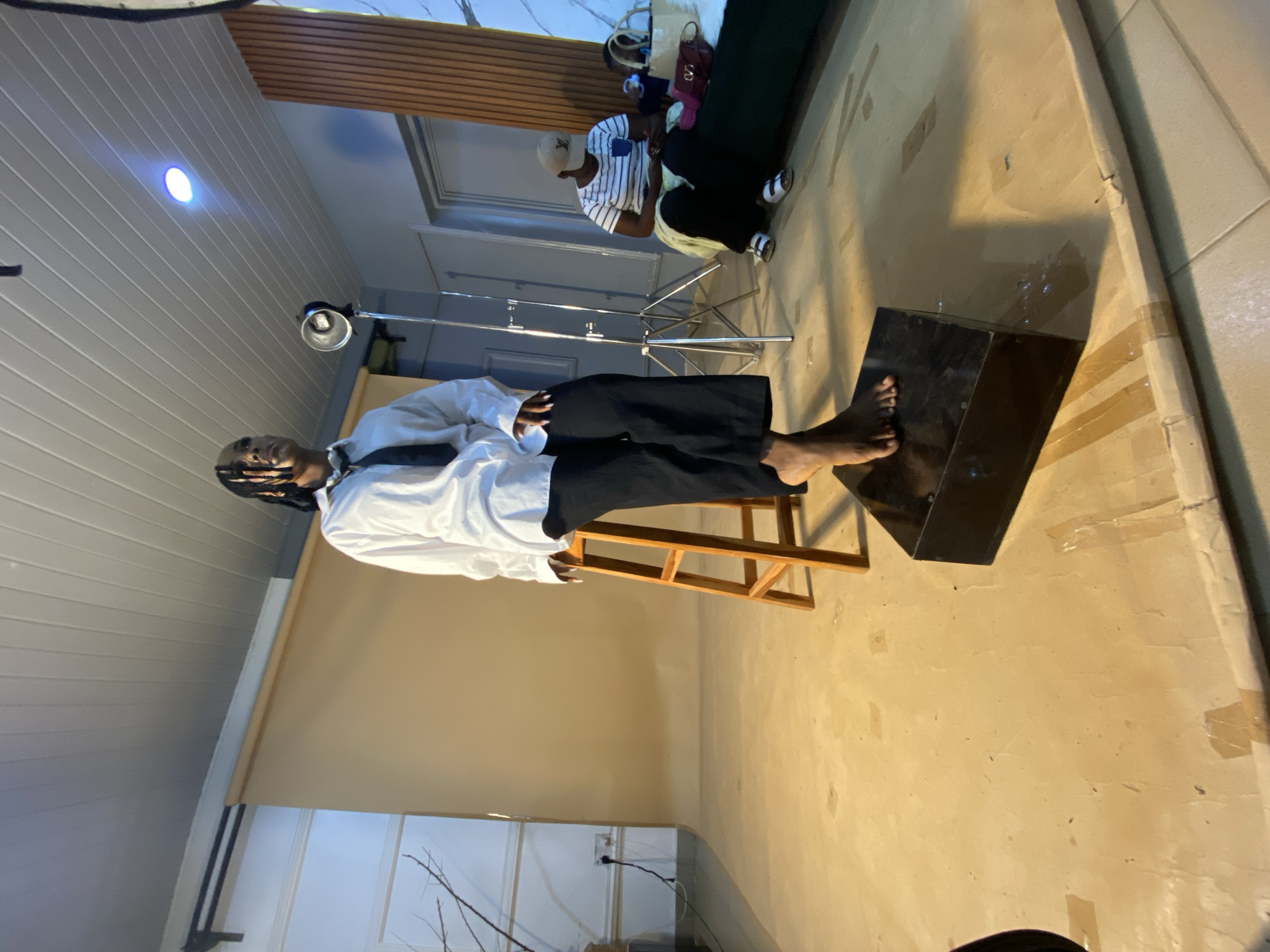 The image depicts an indoor photography studio setup in Bukuru, Nigeria. A person, likely a model, is seated on a wooden stool against a tan backdrop. They are wearing a white button-down shirt and black pants, with a black tie. Their right foot is extended and resting on a dark rectangular prop positioned on the floor. Another individual, wearing a striped shirt and a baseball cap, is seated on a green couch in the background, looking at a mobile device. Several studio lights and equipment are visible, along with some bags and personal items on the couch. The floor appears to be a smooth, light-colored surface taped in places, possibly to secure backdrops or for stage marking. The overall environment suggests a professional or semi-professional photography session.