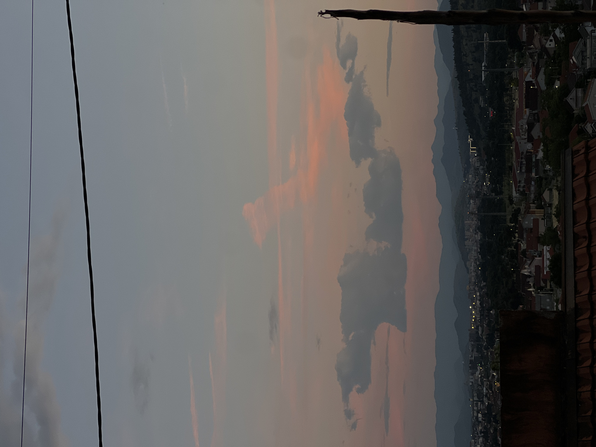 The image shows a sunset scene over Podgorica, Montenegro. The sky is filled with clouds and orange hues. In the distance, a mountain range is visible. The city below has rows of houses with red-tiled roofs and some streetlights. A branch is visible in the upper right. There is a blurry black cable visible on the left side.