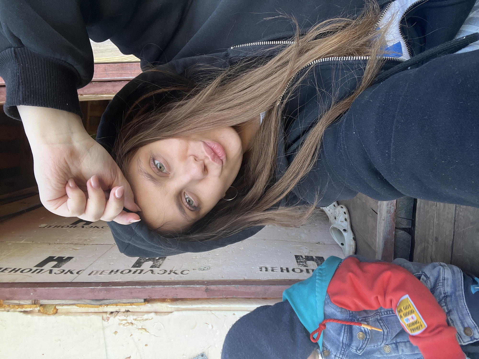 A female individual is depicted in a selfie, positioned in a way that her face is partially obscured by the hood of a black zippered hoodie. The background consists of what appears to be construction or renovation materials, including insulation panels with Cyrillic text (ОЛЬХОВКА ПЕНОПЛЭКС). A denim jacket with a red sleeve is present, featuring a patch with text. A white Croc sandal is visible. The location is presumed to be in Ольховка, Russia, based on the Cyrillic text on the insulation panels.