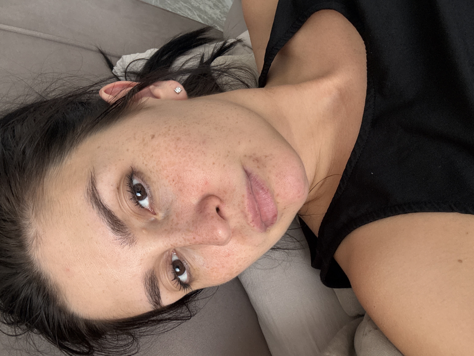 This is a close-up, high-angle selfie of a young woman with dark hair, lying down and looking at the camera. She has fair skin with freckles across her nose and cheeks. Her eyes are brown, and she is wearing minimal makeup. She has a diamond stud earring in her right ear. She is wearing a black tank top. The background appears to be a bed with light-colored pillows and a grey headboard. The image was taken indoors.