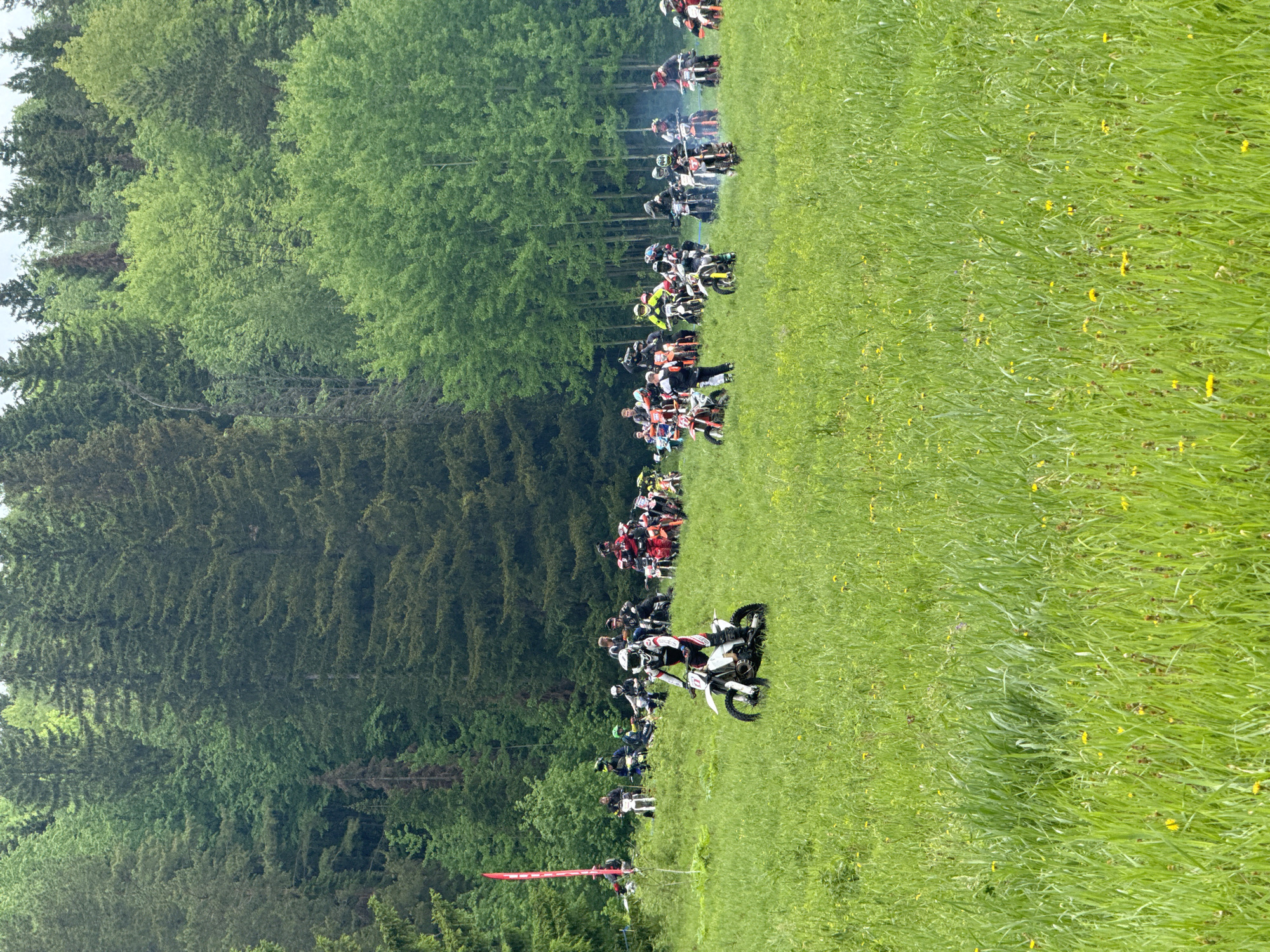 The image shows a group of motorcyclists on a grassy field in Чёрная, Russia. The scene suggests a race or event with a line of motorcycles waiting for the start, near a forest. There are people on the motorcycles and others standing around. A red flag stands prominently at the starting line, signaling the start or a marker. The field has long green grass with some yellow flowers. The left side of the image is bordered by a dense green forest, adding to the rural setting.