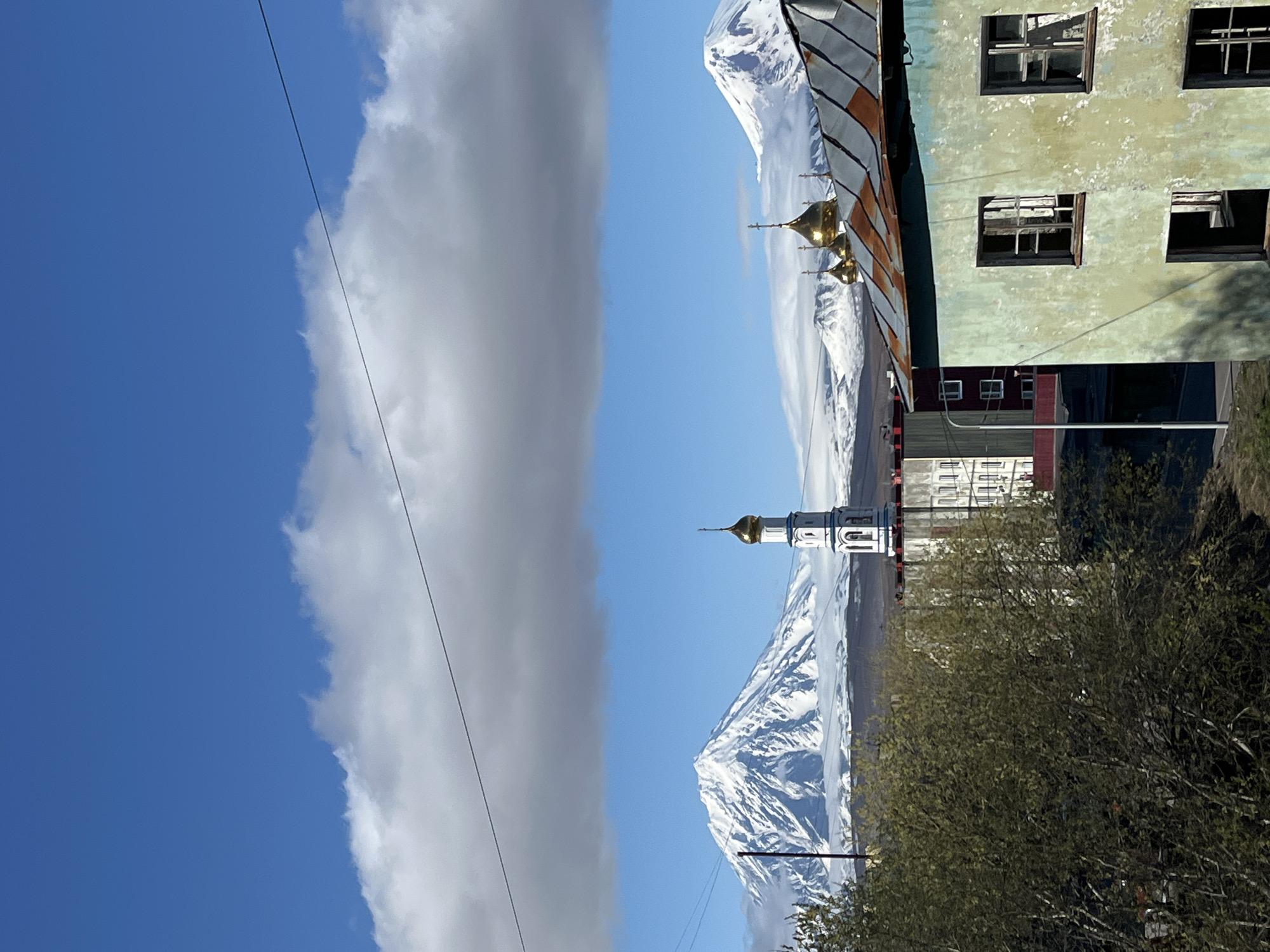 The image shows a view of the city of Petropavlovsk-Kamchatsky, Russia, divided vertically. The left side features a blue sky with a large, gray cloud. The right side depicts a landscape with a snow-covered mountain in the background, partially obscured by a cloudy sky. In front of the mountain is a white church with a golden dome. The foreground contains a building with faded green paint and broken windows. A bush or tree is in the lower right corner of the image. Utility lines are visible in the sky.
