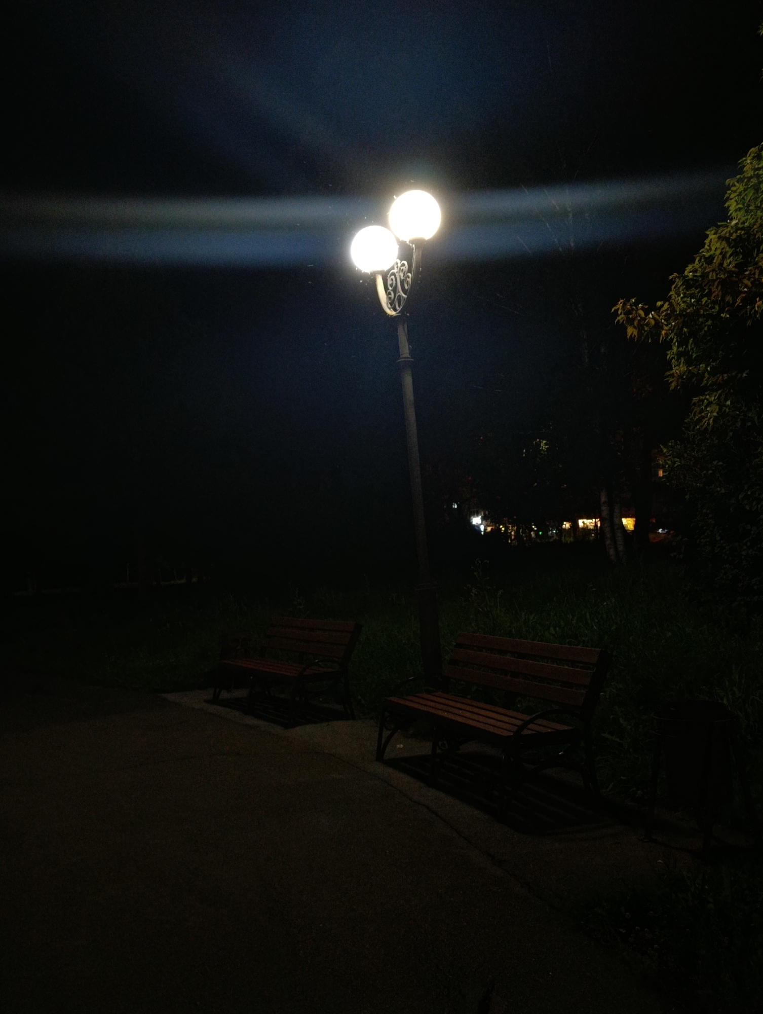 This is a nighttime photograph taken in Abakan, Russia, likely in a park or public area. The scene is dimly lit by a street lamp with two globular lights. The lamp post has decorative metalwork. Two empty wooden benches with metal frames are situated on a paved pathway. Beyond the pathway, tall grass and trees are visible, with some distant lights suggesting buildings or other structures. The sky is dark, with a visible streak of light emanating from the street lamp. The overall impression is one of quiet solitude in a public space after dark.