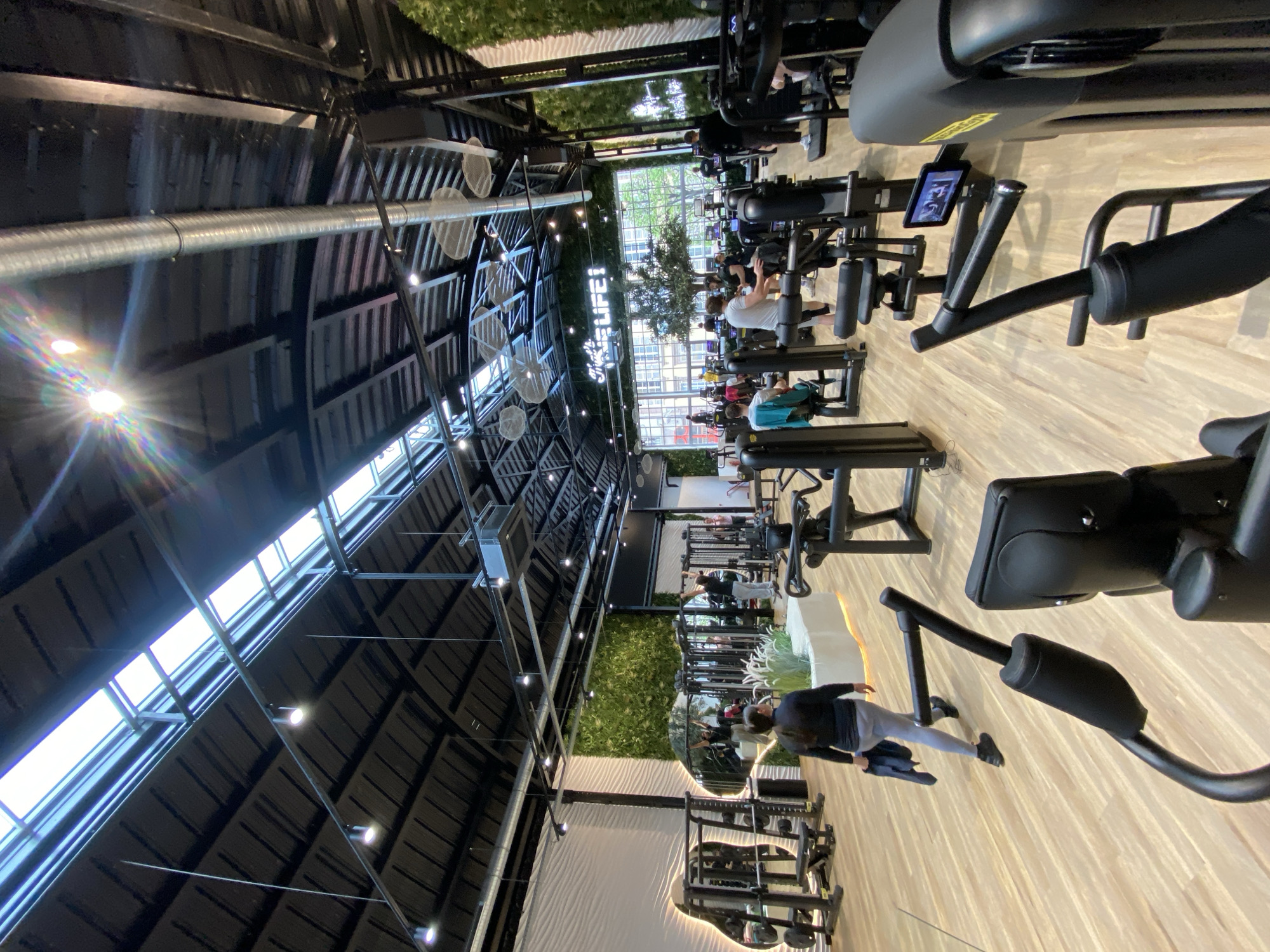 The image shows the interior of a well-lit gym, likely in Strasbourg, France. The gym features rows of modern exercise machines. Several people are exercising on the equipment. The ceiling has a black metal structure with skylights, and there are artificial plants along some of the walls. The floor is a light wood color. There are hanging light fixtures and a large ventilation system running along the ceiling.