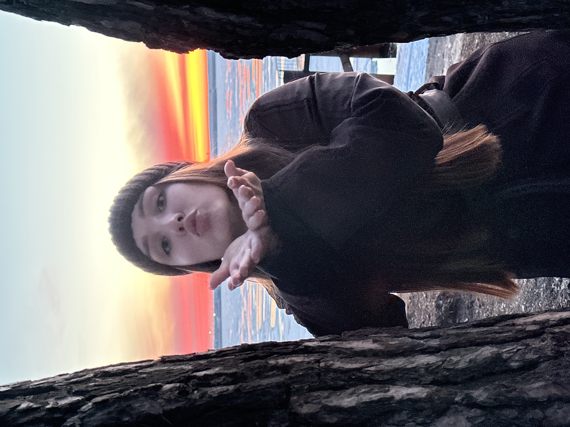 A young woman with light skin and long brown hair is seen wearing a dark beanie and jacket. Her hands are raised near her face, her mouth pursed in a kiss. She is standing behind a dark, rough tree trunk, looking through the space between it and another dark, blurred object above.
Behind her, a body of water reflects a colorful sunset, with hues of yellow, orange, and red. In the distance, the skyline is visible. Given the city name Chaikovsky, Russia, it's plausible the body of water is the Kama Reservoir. A pier structure is partially visible in the distance. The scene appears to be outside, at twilight or dawn, in a natural or semi-natural setting near the waterfront.