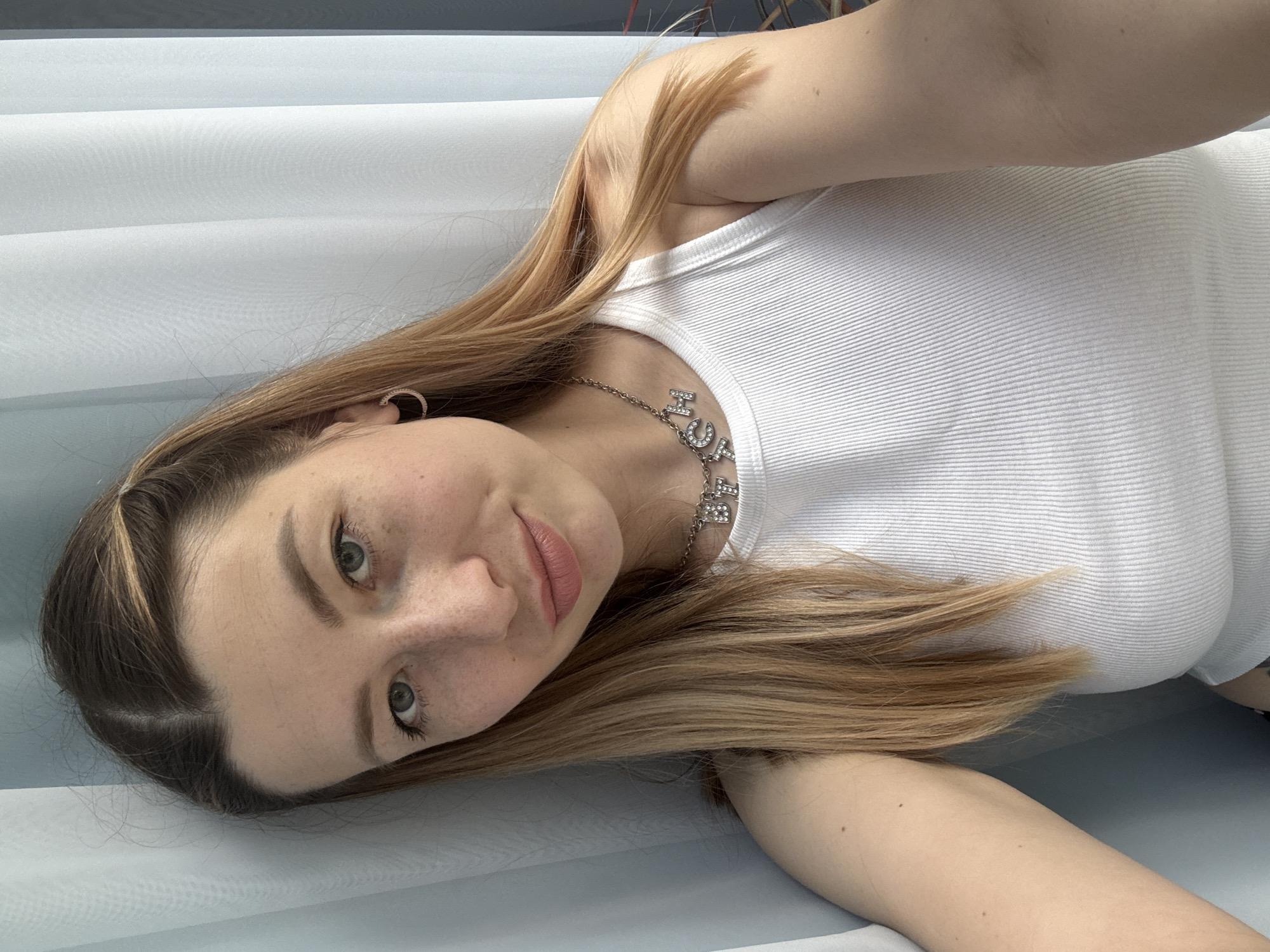 A portrait-style photograph depicts a young Caucasian woman indoors, rotated 90 degrees clockwise, making her appear to be lying horizontally. She is positioned with her head tilted, looking directly at the camera with a calm and gentle expression, featuring a slight smile.
The woman has long, light brown or blonde hair with some darker roots near the part. Her face is adorned with light freckles, and she has light-colored eyes with subtle eye makeup and natural-looking pink lipstick. She wears a simple white ribbed tank top. A silver chain necklace with letter charms is visible around her neck, with the letters "HOC" clearly discernible. A small, sparkling hoop earring is visible on her left ear.
The background consists of a light blue-grey surface or wall, exhibiting subtle horizontal lines, possibly indicating blinds or a textured fabric. The lighting is soft and even, suggesting an indoor setting during the daytime. The overall mood of the image is relaxed and serene. The location context provided is Moscow, Russia.
