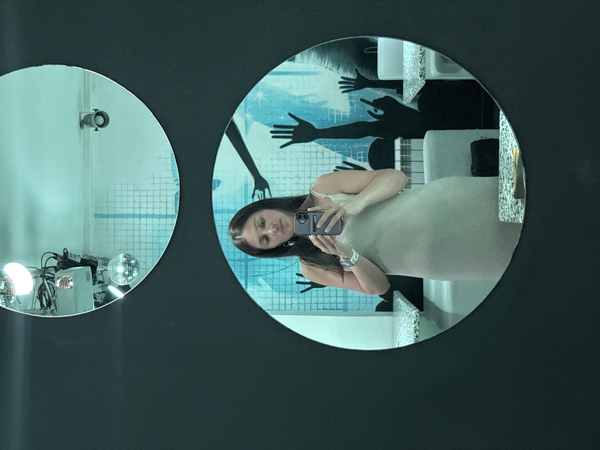 A person is taking a selfie in a round mirror. In the mirror, a person with dark hair is holding up a cell phone. The background shows decorations including hands and tiles. To the left, a second round mirror reflects equipment, including disco balls. The background color is black. There is no location information available to identify whether the location is Chaikovsky, Russia.
