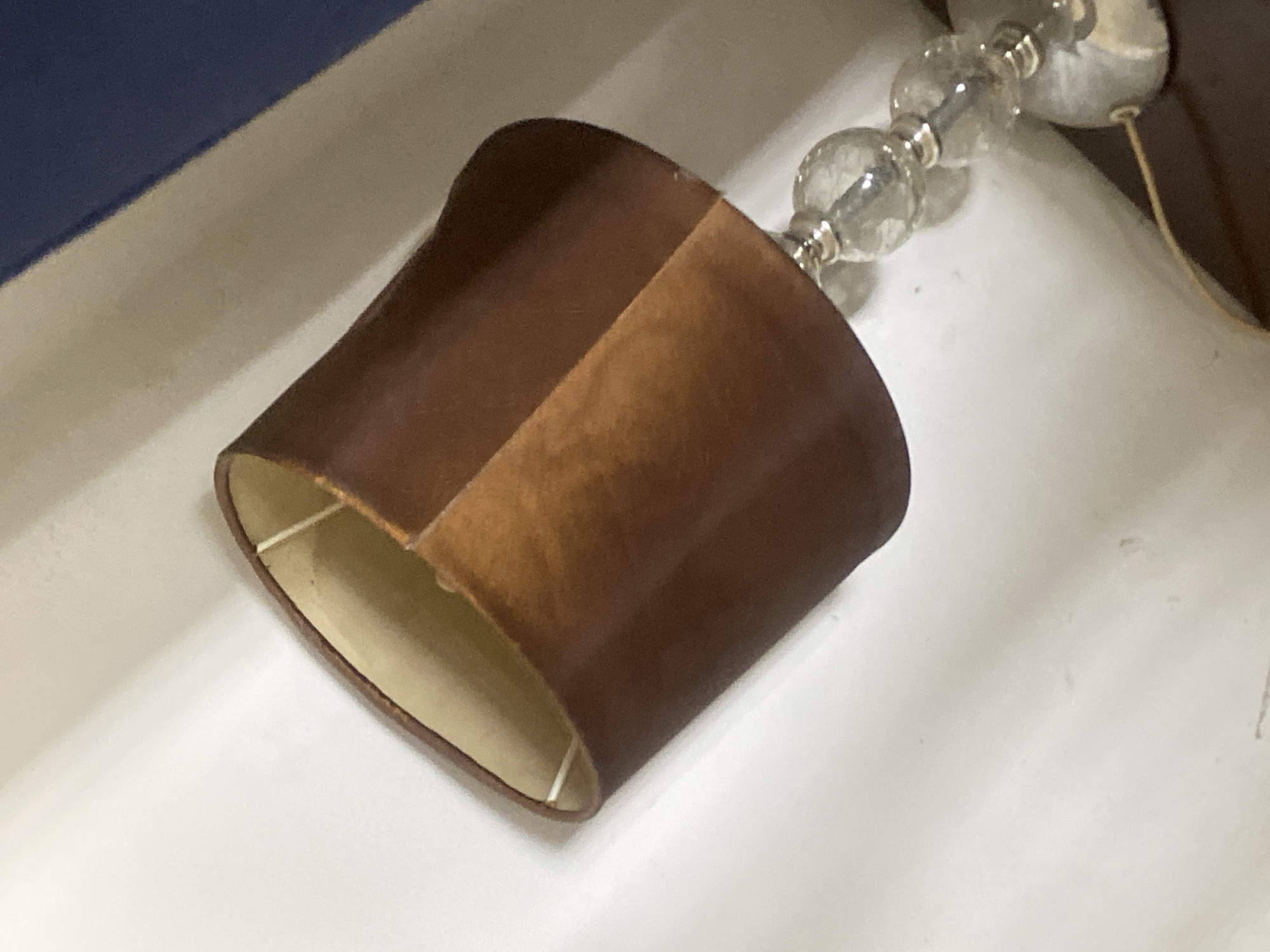 A close-up shot shows a brown lampshade with a white interior resting on a white surface. The lampshade is slightly angled. Attached to the top right of the lampshade are clear glass beads with silver accents. The top left of the frame includes a white surface and a navy blue object. The scene appears indoors under diffused lighting. The image does not contain information to determine the location in the city of Abuja, Nigeria.