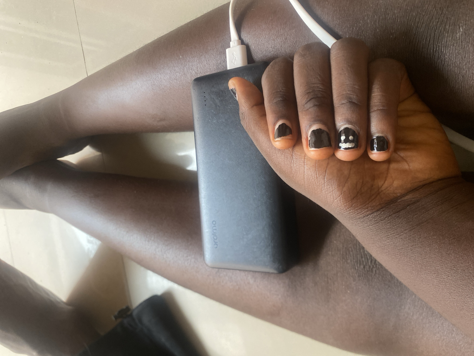 A close-up shot shows a person's hand resting on a dark gray power bank, which is plugged into a white cable. The hand has dark skin and is adorned with black nail polish. One nail on the index finger is painted with a simple smiley face. The power bank has the "oraimo" logo printed on its side. The scene appears to be indoors, on a tiled floor, with the lighting suggesting daytime. The person's legs are also visible, with one leg bent and resting on the other. The overall impression is casual and relaxed, possibly indicating the person is charging their device while taking a break. The location context is Wauro Leggal, Nigeria.