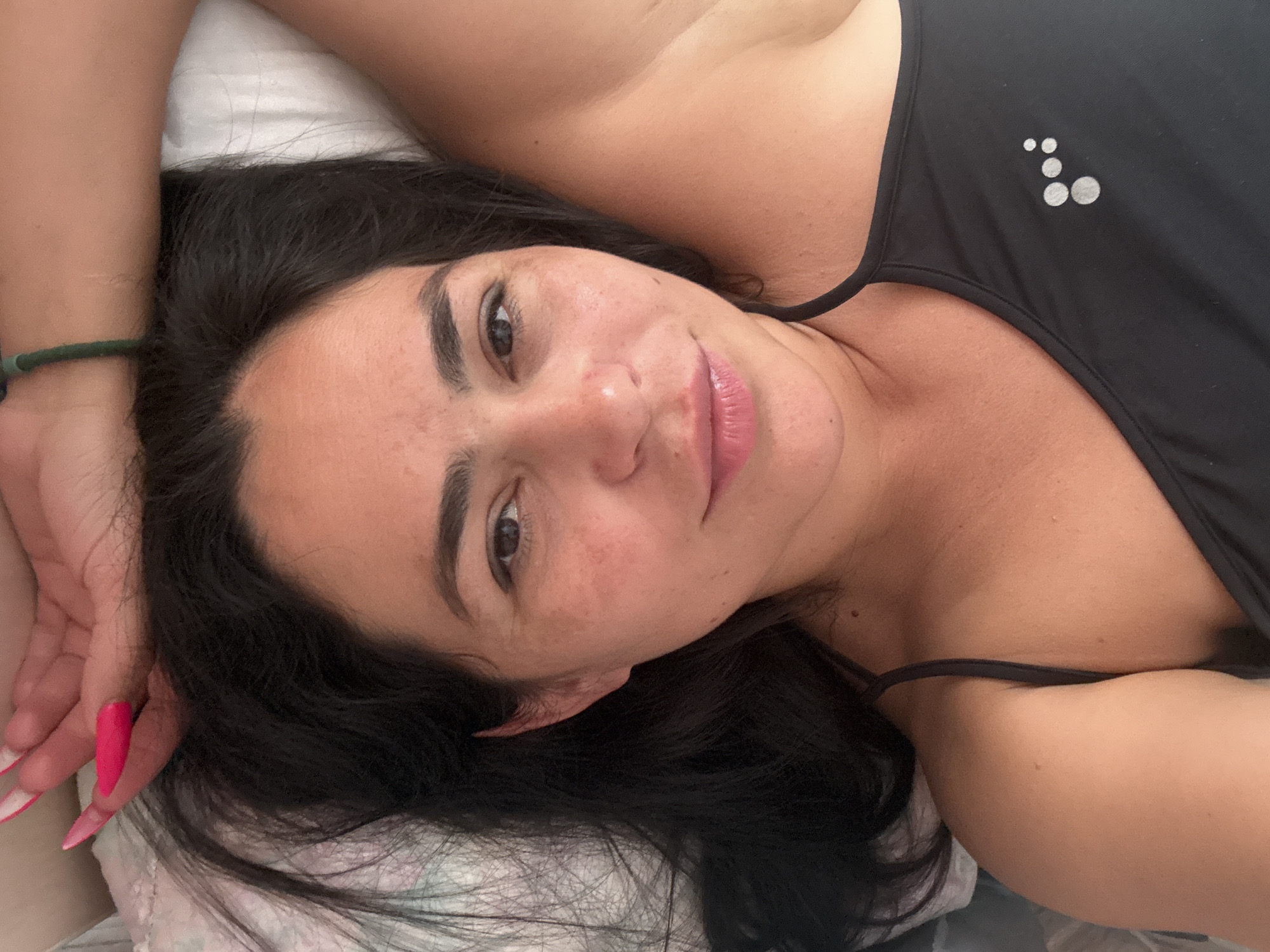 A woman lies on a bed. She has dark hair, and is wearing a black tank top. Her left arm is bent with her wrist above her head. Her nails are bright pink. She is likely indoors, on a pillow, and looking at the camera. Location cannot be determined.