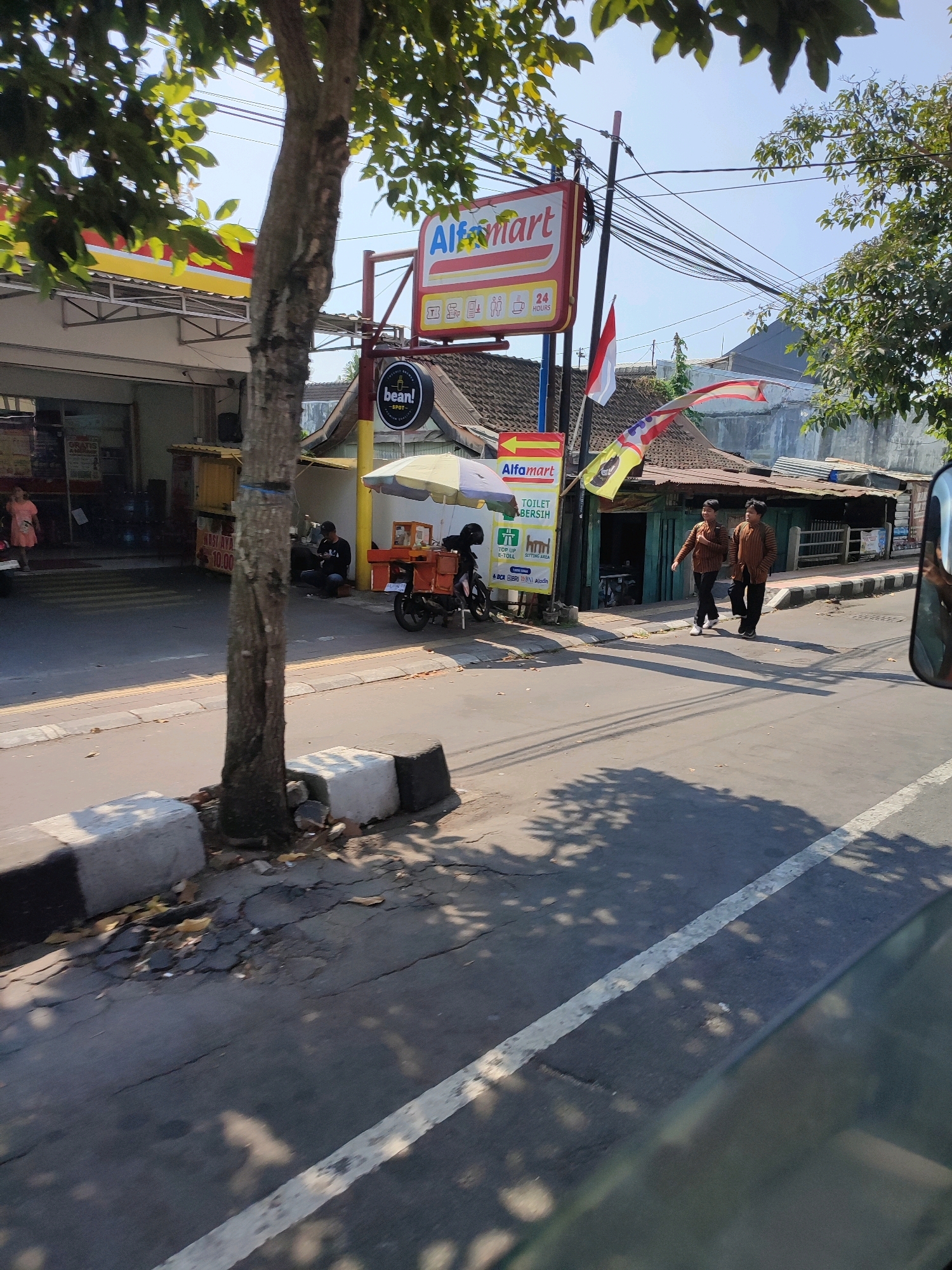 The image depicts a street scene in Magelang, Indonesia, with a prominent Alfamart store on the left. A young woman in a pink dress is seen near the entrance of the store. In front of the store, there is a vendor cart with a yellow umbrella, and a person is seated behind it. A motorcycle is parked next to the cart. Two men, dressed in identical brown patterned shirts and dark pants, are walking on the sidewalk. Several Indonesian flags are visible, one of which is a red and white flag on a pole. The street is paved and shows some signs of wear and tear, with cracks and potholes in places. Trees line the street, casting shadows on the pavement. Electrical wires are strung across poles in the background. The sky is clear and blue.