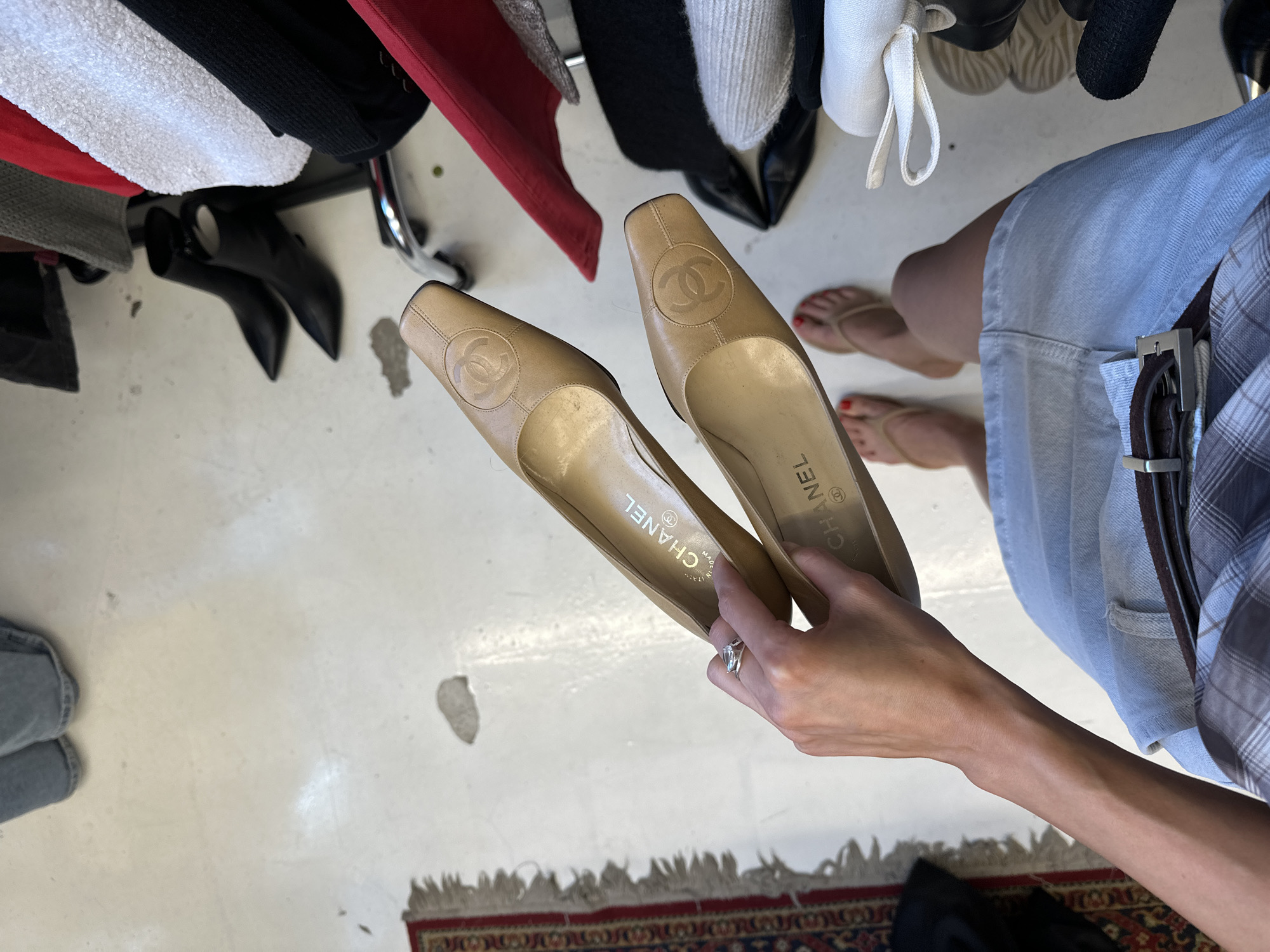 The image depicts a pair of beige leather Chanel pumps being held by a woman's hands. The shoes have a square toe and the signature Chanel CC logo embossed on the vamp. The woman is wearing a light blue denim skirt and a plaid shirt. Her feet are bare, with red nail polish on her toenails. She is wearing a ring on her left ring finger. In the background, there are clothing items hanging on a rack, including a white textured top, a black garment, and a red garment. The scene appears to be indoors, on a light-colored floor. There is no indication of the location being Moscow, Russia.