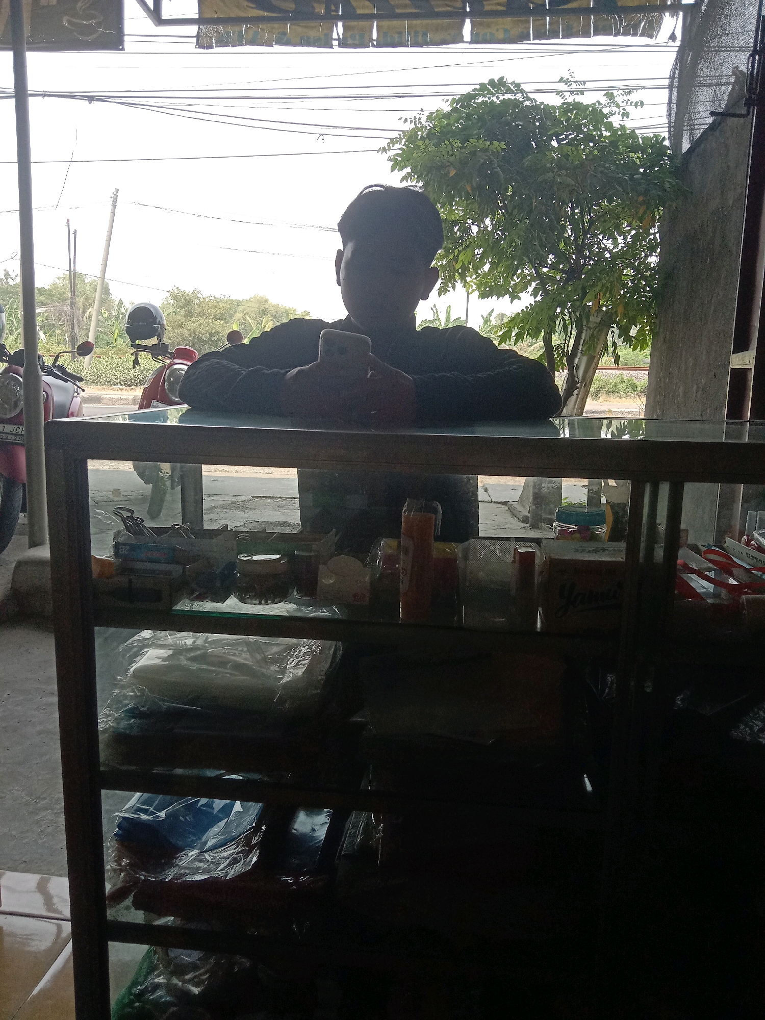 A young man, appearing to be in his late teens or early twenties, stands behind a glass-topped display counter. He is silhouetted against the bright outdoor light, holding a smartphone in his hands, and looking down at it. The counter contains various items, including packaged goods and what appear to be cleaning supplies. Beyond the counter, a red and a white motorcycle are visible parked outside, along with greenery and trees. The scene appears to be a small shop or stall, possibly in a rural or semi-urban area. The man's posture suggests he is engaged with his phone, oblivious to his surroundings. The overall lighting is bright, with the outdoor environment significantly lighter than the interior of the shop.