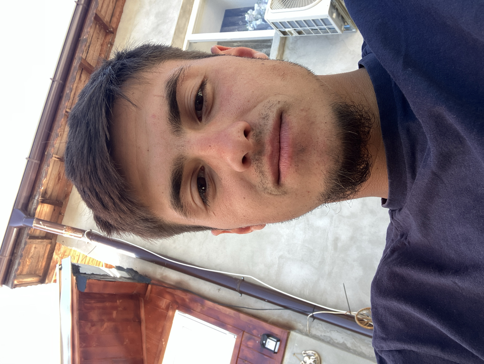 A young man is taking a selfie in Komuniga, Bulgaria. He has dark hair and a stubbly beard, and is wearing a dark blue t-shirt. Behind him is a building with a wooden roof and a white wall. An air conditioning unit is visible near the top of the frame.