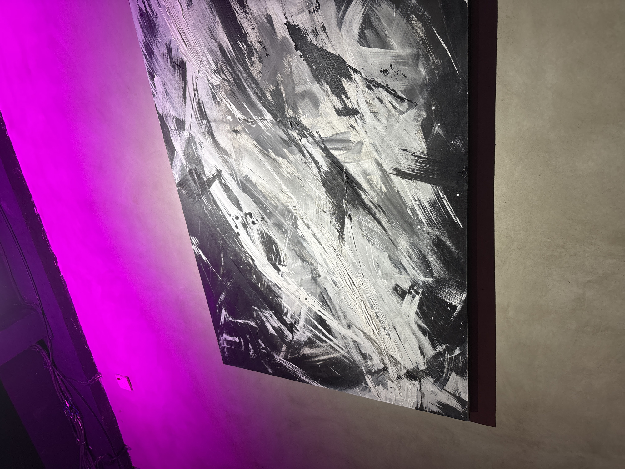 A medium shot captures an abstract black and white painting hanging on a light-colored wall, located in Kuta Selatan, Indonesia. The painting features dynamic, bold brushstrokes of white and shades of gray against a black background. The painting is framed with a dark, possibly burgundy, border. A section of the adjacent wall is bathed in bright, neon pink light. Wires are visible on the left side of the image. The overall setting appears to be indoors, possibly an art gallery or modern interior space.