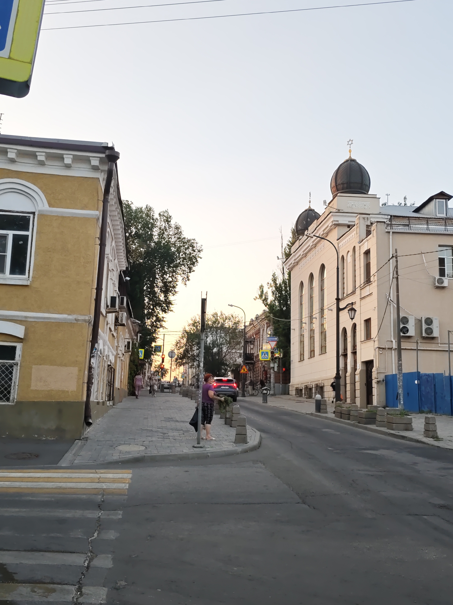 A view down a city street in Rostov-on-Don, Russia, with buildings on both sides. The sky is clear and pale. On the left, a yellow building with a white trim and arched windows is visible. On the right, a cream-colored building with a dark dome and a Star of David on top is prominent. The street is paved with asphalt and there is a pedestrian crossing in the foreground. People are walking on the sidewalk, and a car is driving on the street. The atmosphere suggests it is late afternoon or early evening.