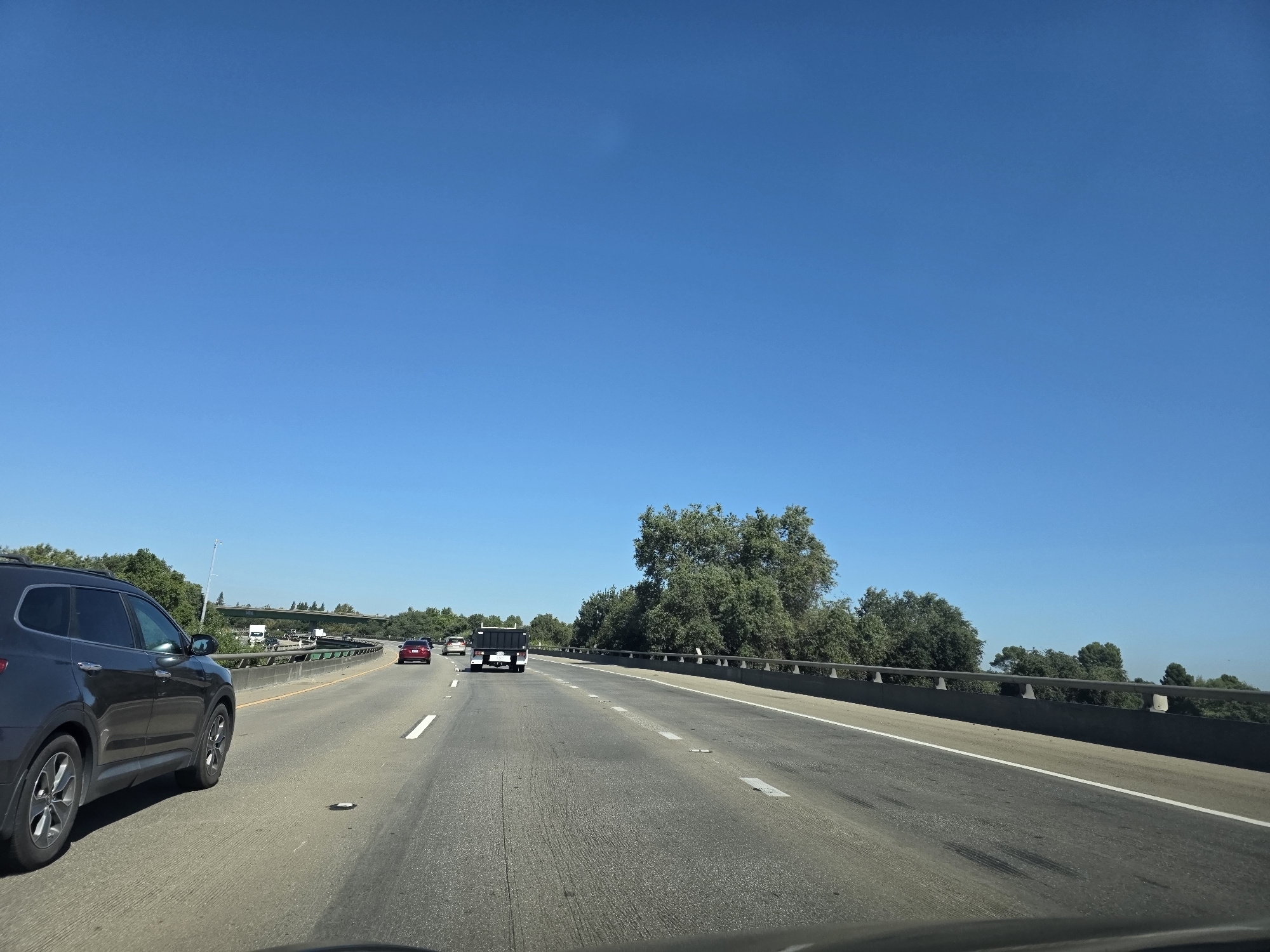 The image depicts a view from inside a vehicle traveling on a multi-lane highway under a clear blue sky. The highway appears to be in a suburban or rural area with trees and vegetation lining the right side. On the left, there is a dark-colored SUV in the foreground, and further ahead, other vehicles are visible, including a red sedan and a black utility vehicle. An overpass or bridge structure can be seen in the distance. The road surface shows lane markings and some wear. The overall scene suggests a sunny day with moderate traffic on a highway. Based on the provided information, this scene is located in West Sacramento, USA.