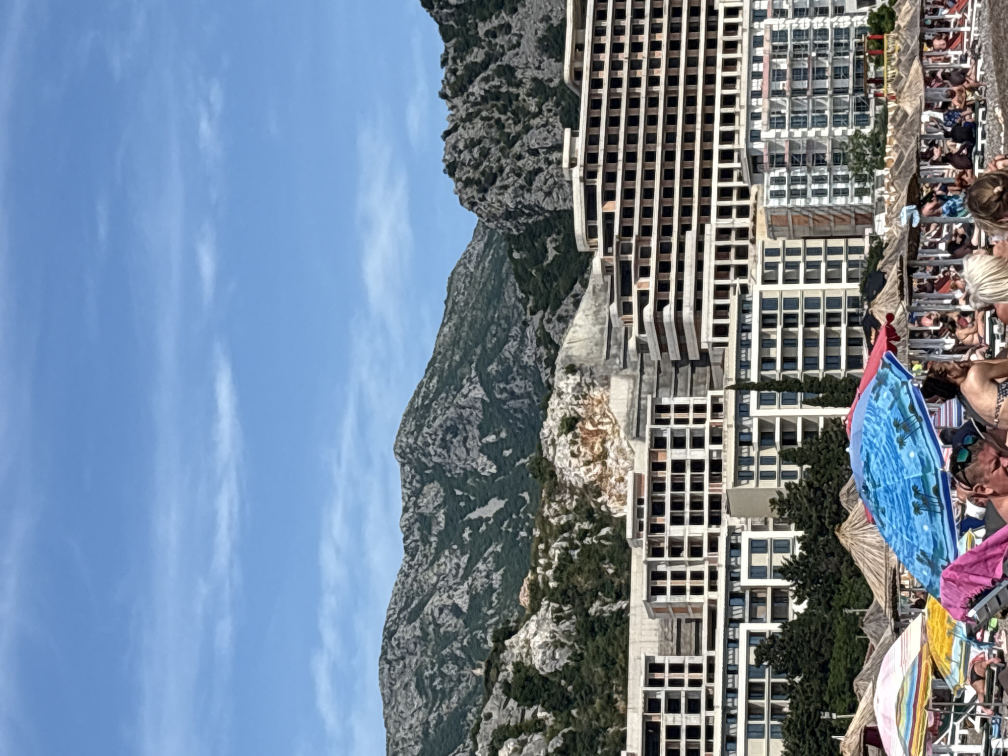 The image displays a daytime scene at a beach in Čanj, Montenegro. The sky is clear blue with sparse, wispy white clouds. A rugged mountain range, characterized by grey rock and green vegetation, dominates the background. On the lower mountain slopes, exposed light-colored rock faces are evident. Directly in front of the mountains, a large complex of multi-story buildings is under construction or partially completed, featuring exposed concrete frameworks and numerous empty window apertures. Some lower levels of these structures exhibit finished facades and windows, interspersed with dark green coniferous trees. The foreground shows a crowded pebble beach occupied by numerous individuals. Many people are seated or reclining on lounge chairs beneath a variety of beach umbrellas, including a prominent blue umbrella with palm tree patterns and a striped multicolored umbrella in the immediate foreground, alongside numerous thatched straw umbrellas extending into the mid-ground. Beachgoers are dressed in swimwear, engaged in passive leisure activities.