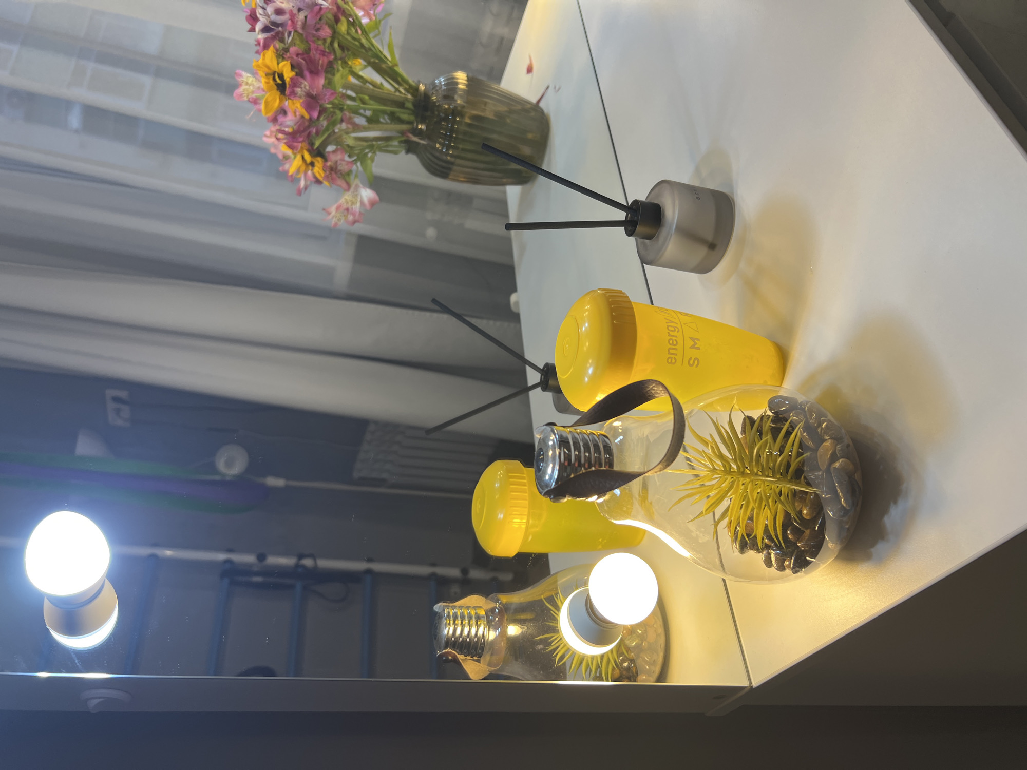 The image shows a vanity table with various items, reflected in a mirror. On the table, there are two decorative glass lightbulb-shaped containers with plants and stones inside, and a third, identical container in the background. There are also two yellow containers, one with the text "energy SMA" visible. A grey glass diffuser with reeds stands to the right. In the mirror's reflection, a bouquet of pink and yellow flowers in a dark green vase is visible, along with sheer white curtains and a window. The mirror also reflects a bright light bulb, a ladder, and some colorful straps. The scene is lit by the bright bulb, creating a warm glow. The overall impression is that of a well-decorated personal space. The image does not provide any information about the city of Sochi, Russia.