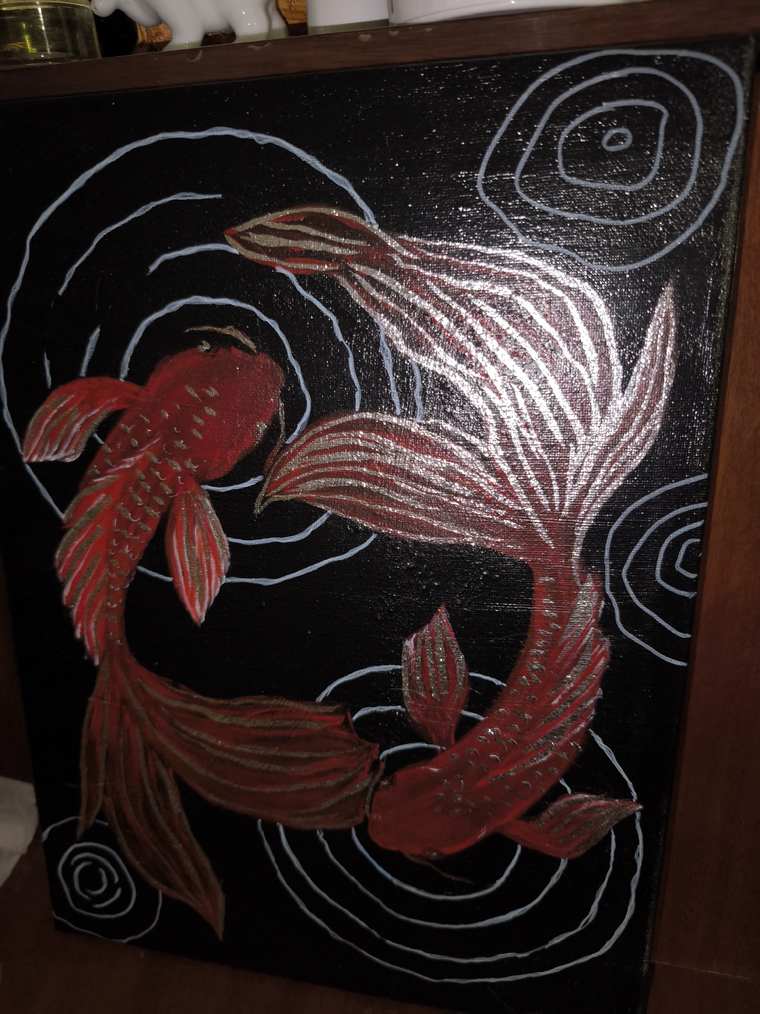 The image displays a black canvas painting of two red koi fish swimming in a circular pattern, symbolizing yin and yang. The fish have metallic silver accents on their fins and tails, and the water is depicted with white swirling lines. The painting is framed by a wooden border. The setting is indoors, with a shelf above the painting holding various objects, including a glass jar and a white figurine. The overall scene appears to be a domestic interior. There is no indication of the city of Bolshaya Martynovka, Russia, in the provided image or its context.