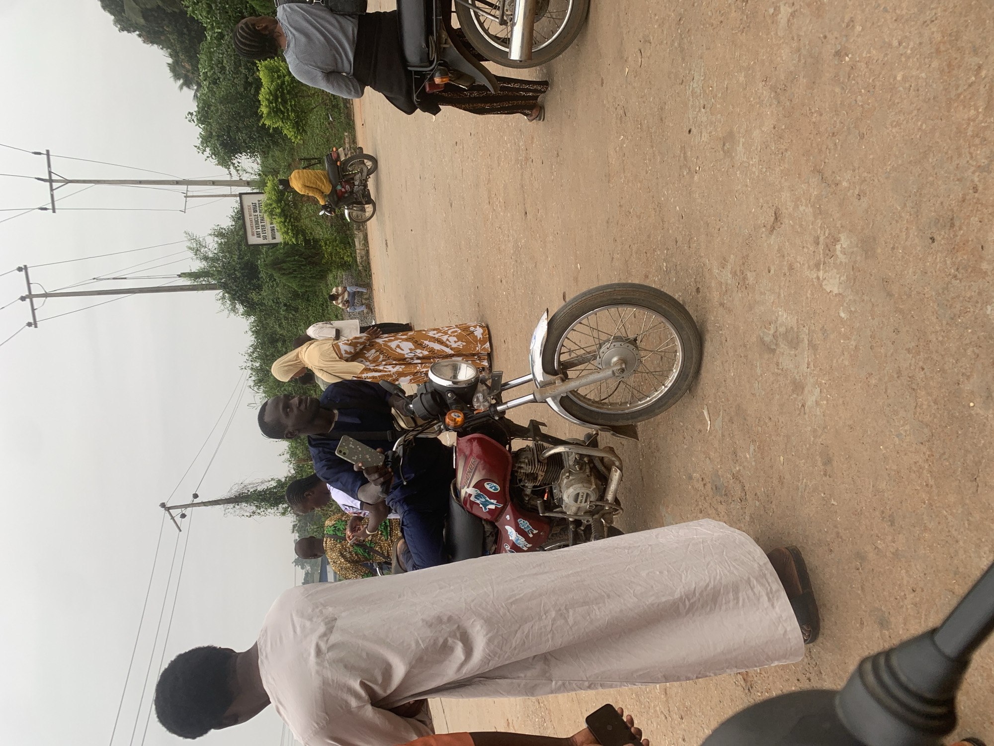The image depicts a street scene in Shagamu, Nigeria, under an overcast sky. In the foreground, a person in a long, light-colored robe stands facing away from the camera, with a dark sandal on their right foot. To their left, a hand holds a dark smartphone. Adjacent to this person is a red motorcycle, with the brand name "BOXER" visible. A man in a dark blue outfit is seated on the motorcycle, holding a smartphone in his right hand. The motorcycle has decorative decals.
Behind them, a woman in a light-colored hijab and a patterned dress stands to the left. Further back, a man in a yellow jacket is seated on another motorcycle, facing left. A woman in a grey top and patterned trousers is visible on the right, also facing left, with a black bag on her shoulder. Several other individuals and motorcycles are present in the background, along with telephone poles and lush green vegetation. The ground is a dusty, light brown color. A white sign with black text is mounted on a pole in the background.