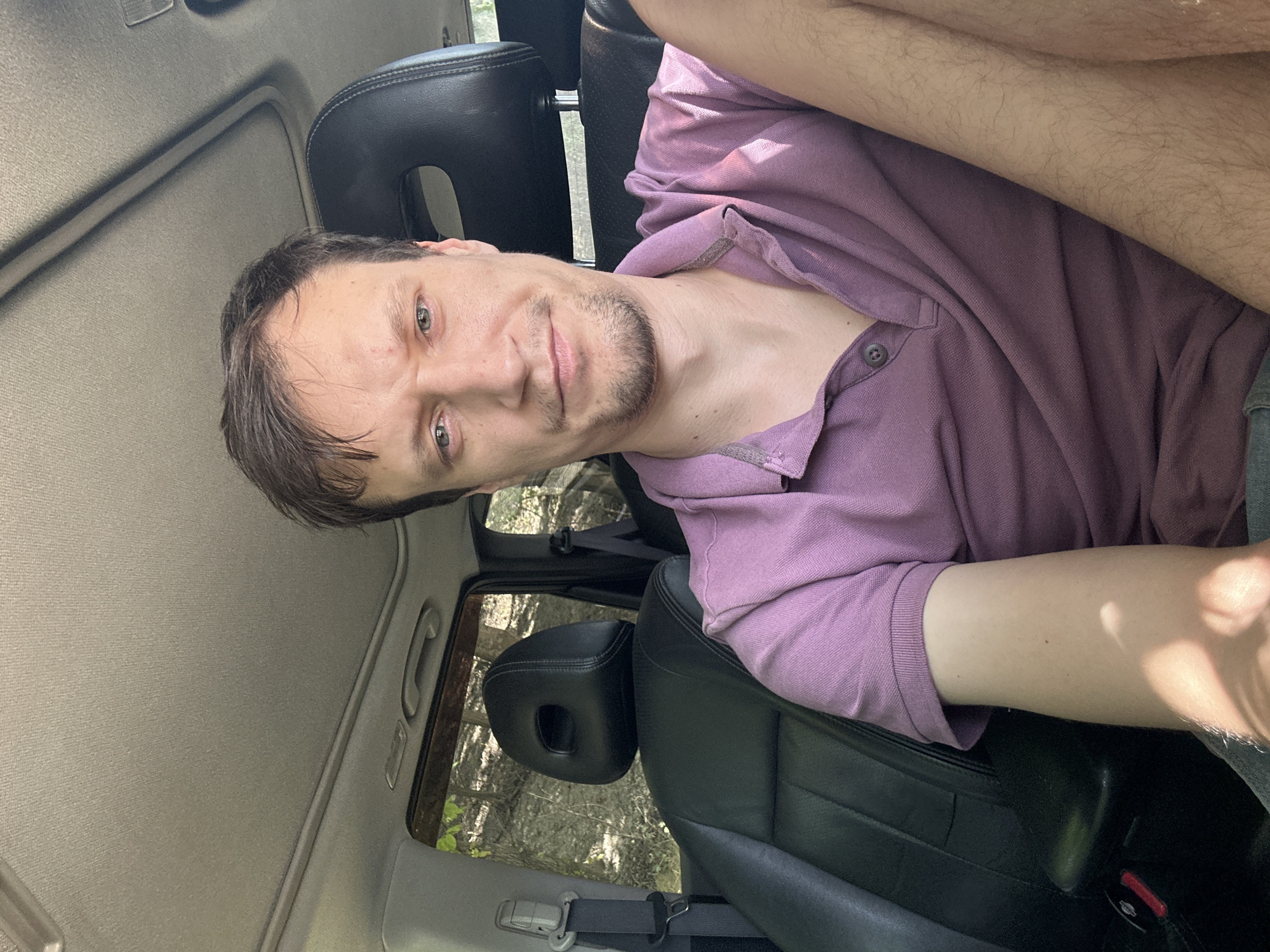 A light-skinned man with short brown hair and light green eyes is seated in the driver's seat of a car. He is wearing a purple polo shirt and has light stubble on his face. The interior of the car is visible, including black leather seats and a sunroof. Outside the car, trees and foliage suggest a rural or wooded setting. The image appears to be a selfie taken by the man. The specific location in the city of Makiv, Ukraine cannot be determined from the image alone.